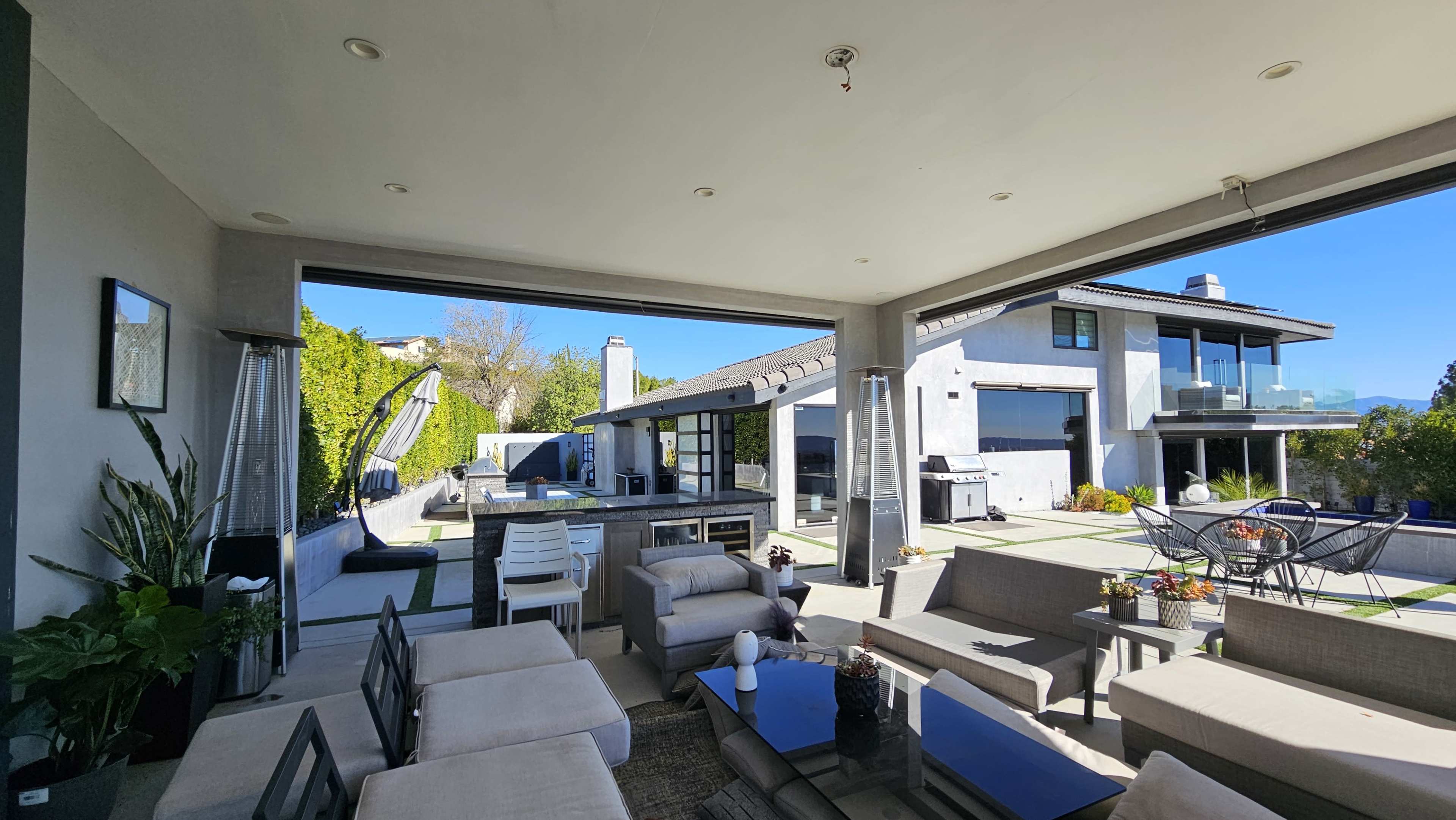 The image showcases a modern outdoor living space with a seating area, dining table, and a view of a contemporary house surrounded by greenery.