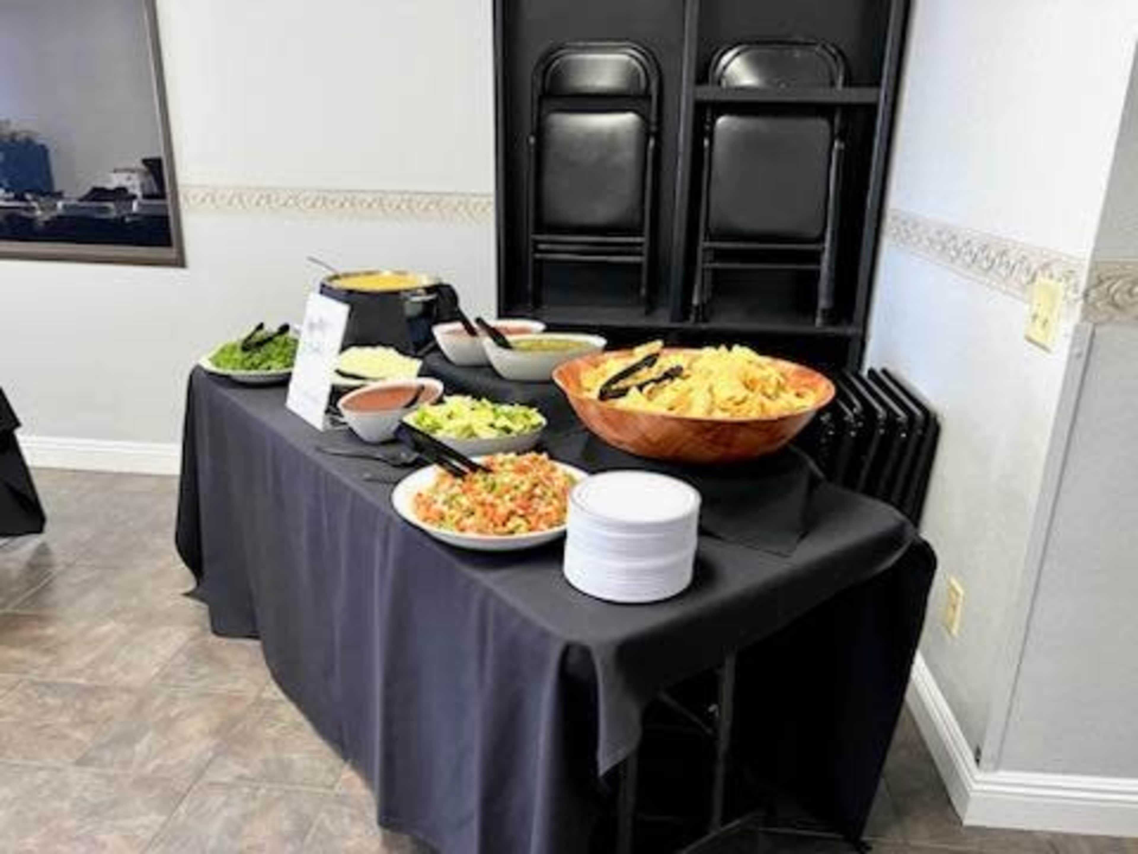 A buffet table is set up with various bowls of salad, chips, and sauces, alongside a stack of white plates, in a room with folding chairs leaning against the wall.