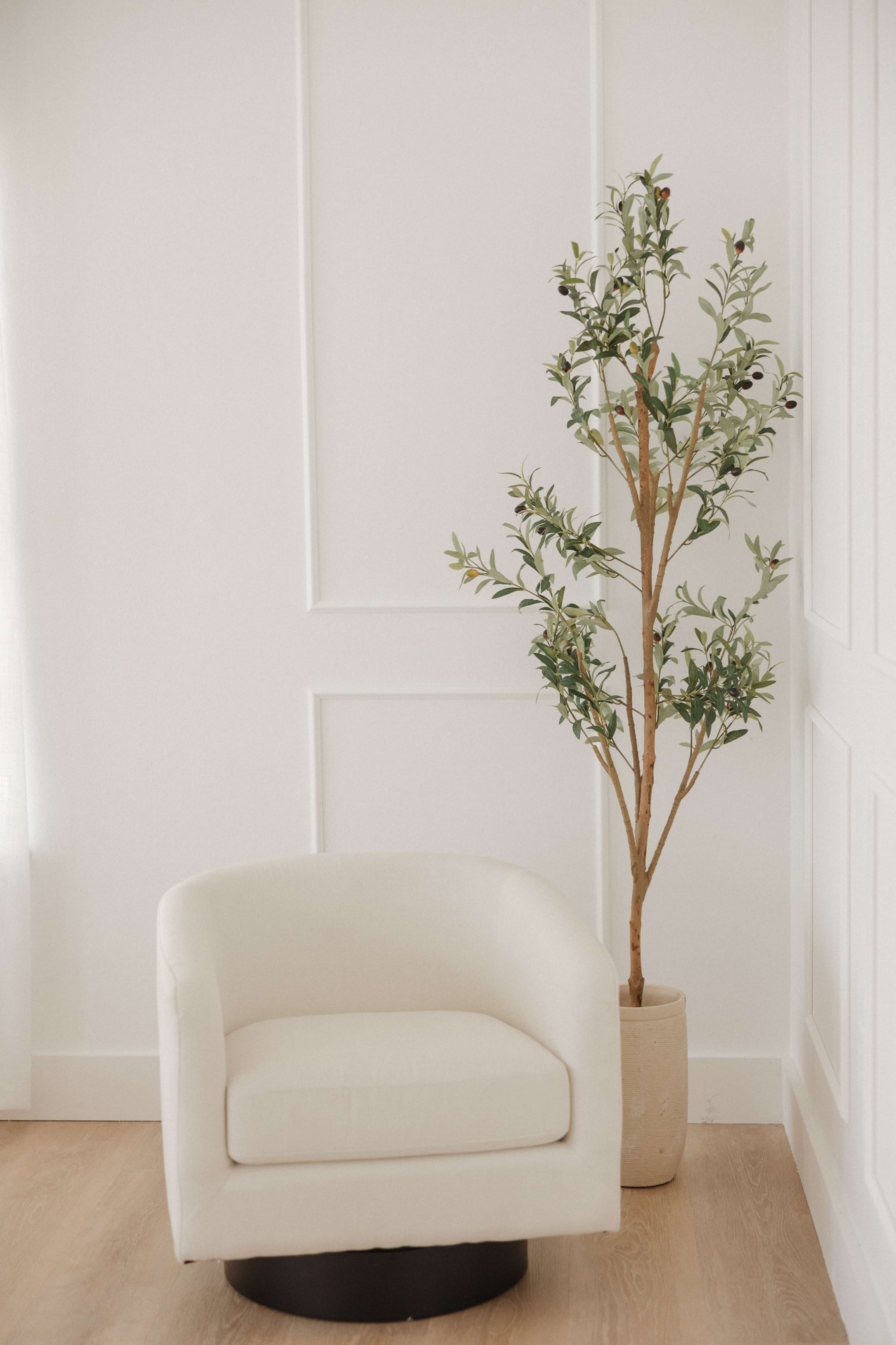 The image features a small, white upholstered chair next to a potted artificial tree against a plain white wall.