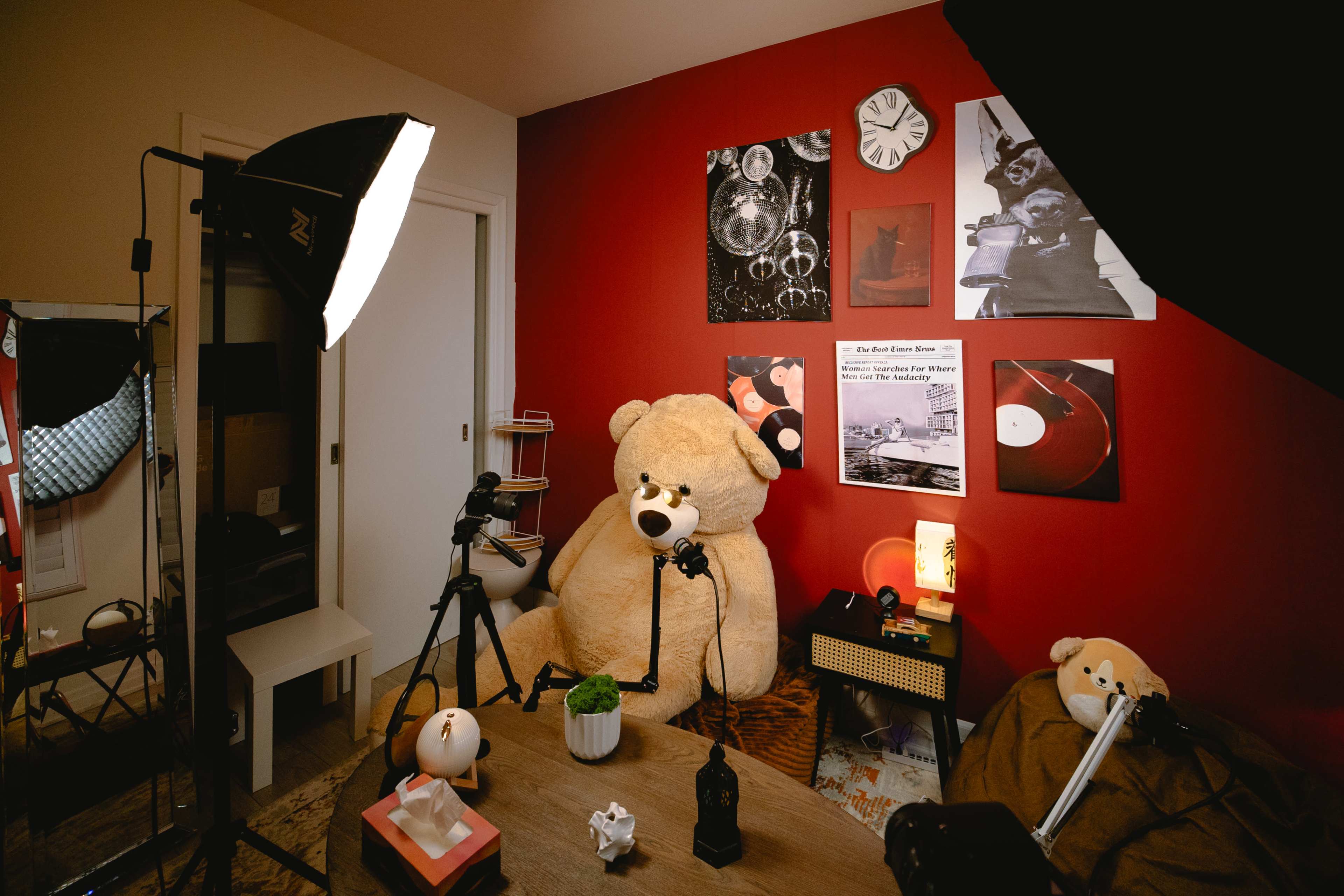 A cozy indoor setup features a large teddy bear sitting in front of a red wall adorned with photographs, lighting equipment, and a wooden table with various decorative items.