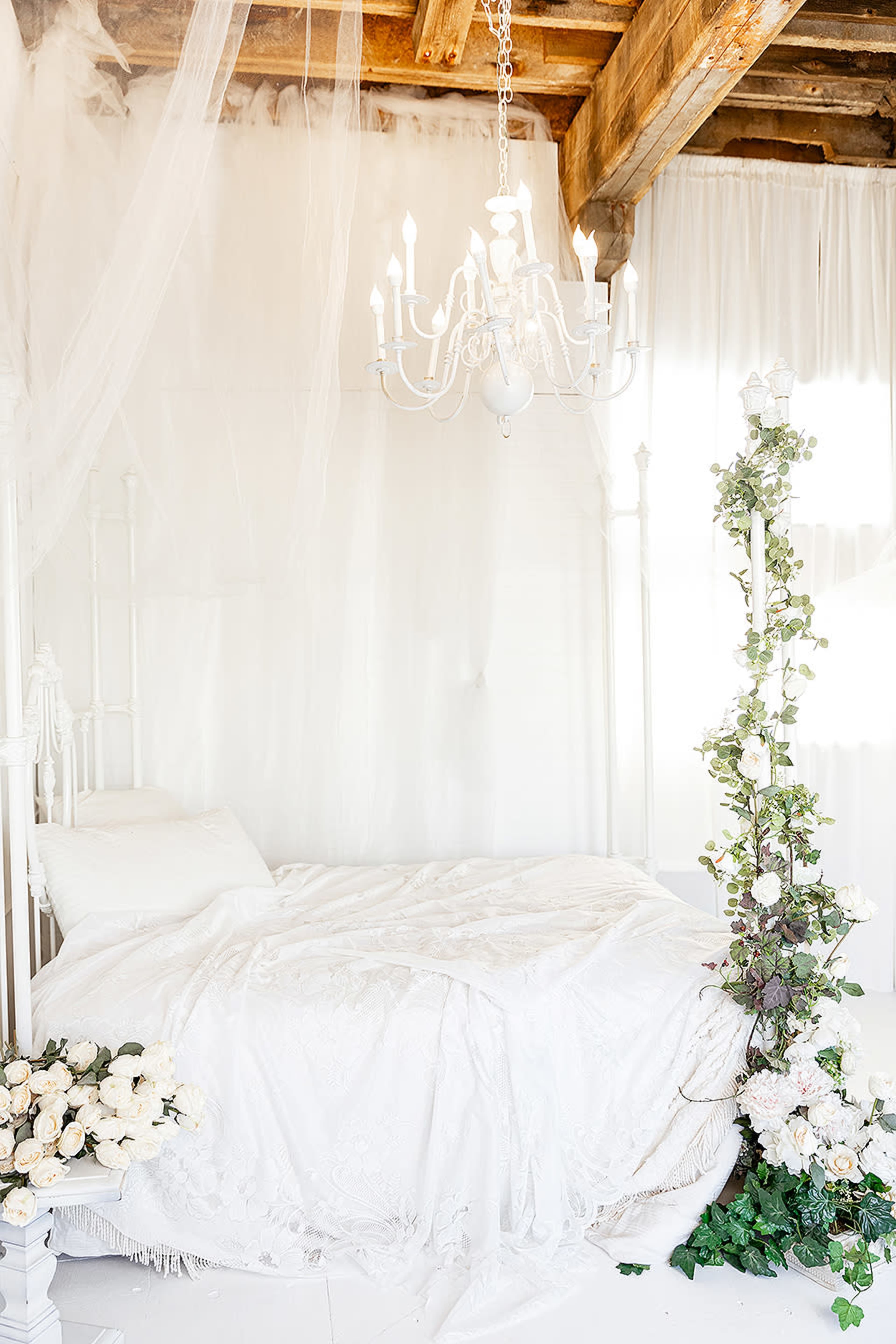 A white canopied bed is adorned with floral arrangements and a chandelier hangs above in a bright, airy room.