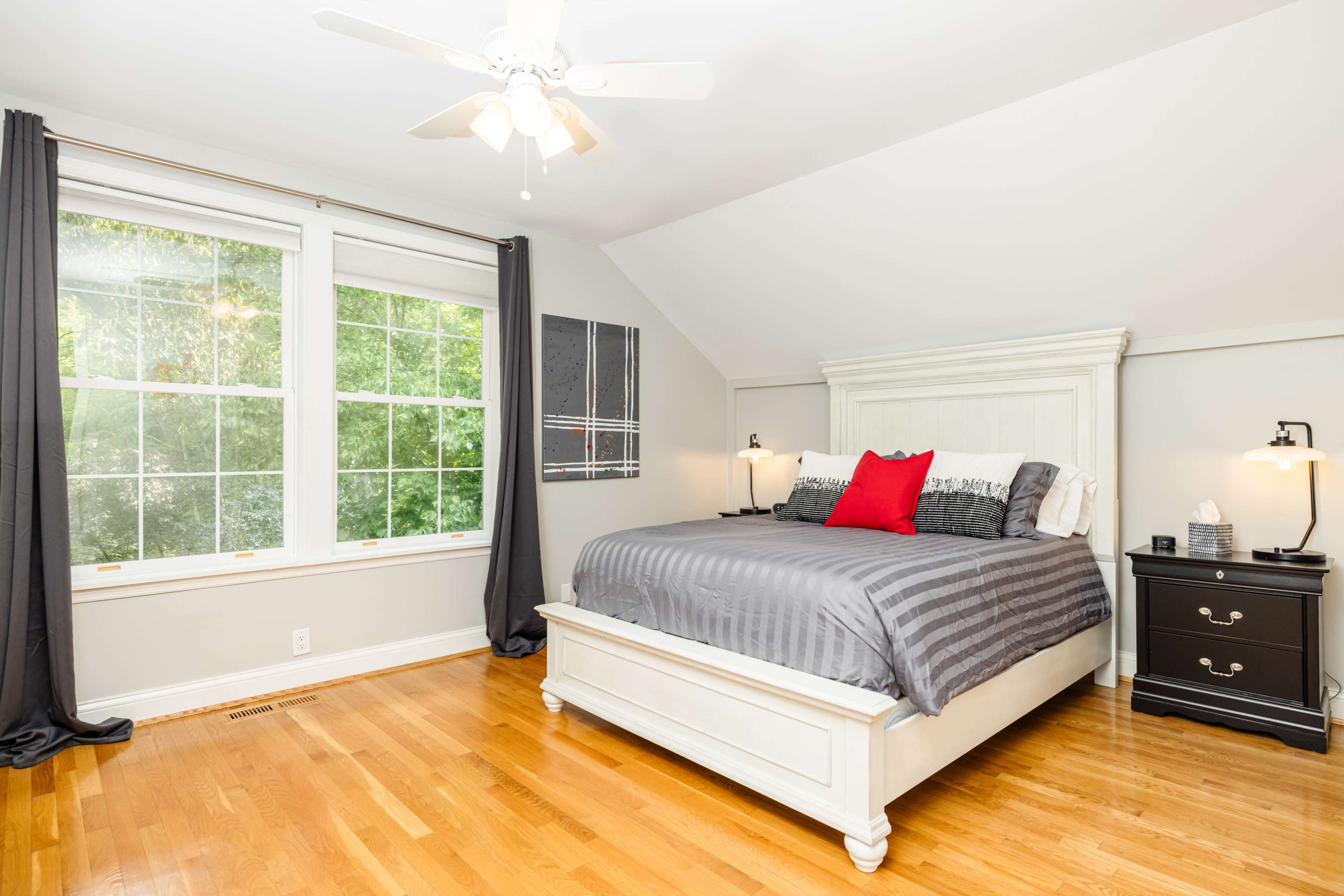 A well-lit bedroom featuring a white bed with gray bedding, a red accent pillow, and a nightstand beside it, along with large windows and hardwood flooring.