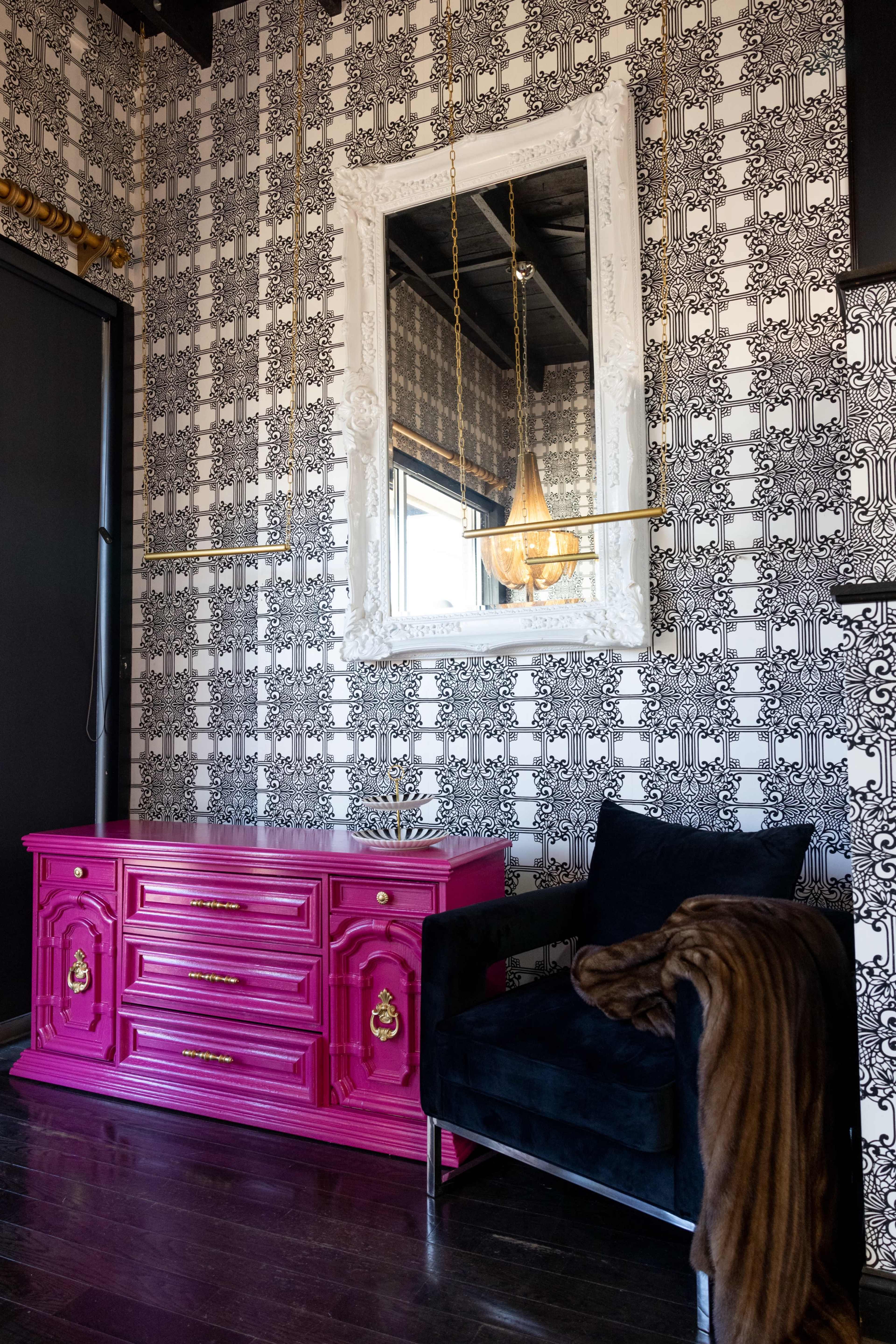 The scene features a pink dresser against a black and white patterned wall, with a large ornate mirror above it and a dark armchair to the side.