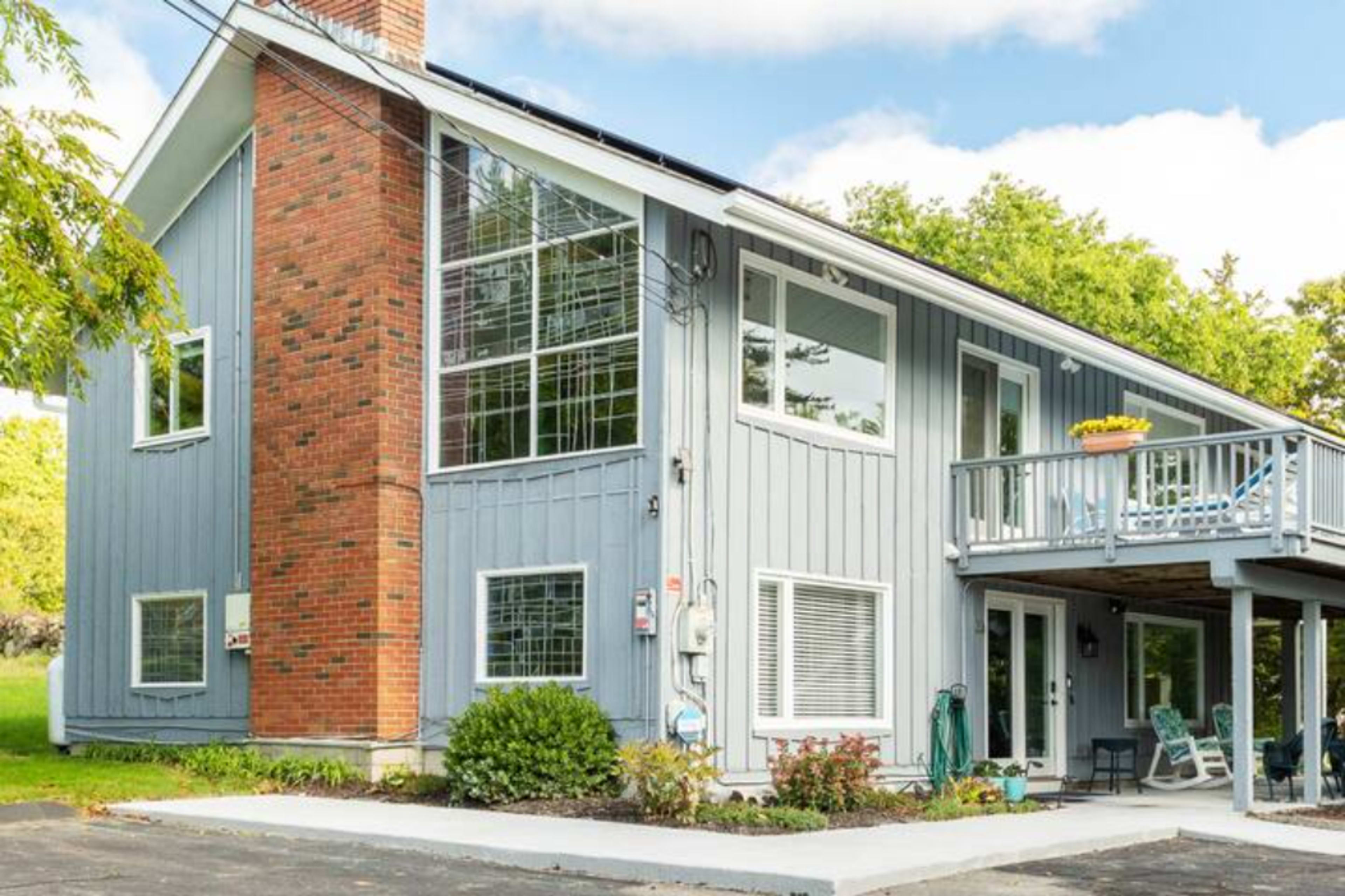 A modern two-story house with a combination of gray siding and red brick, featuring large windows and a front porch with seating.