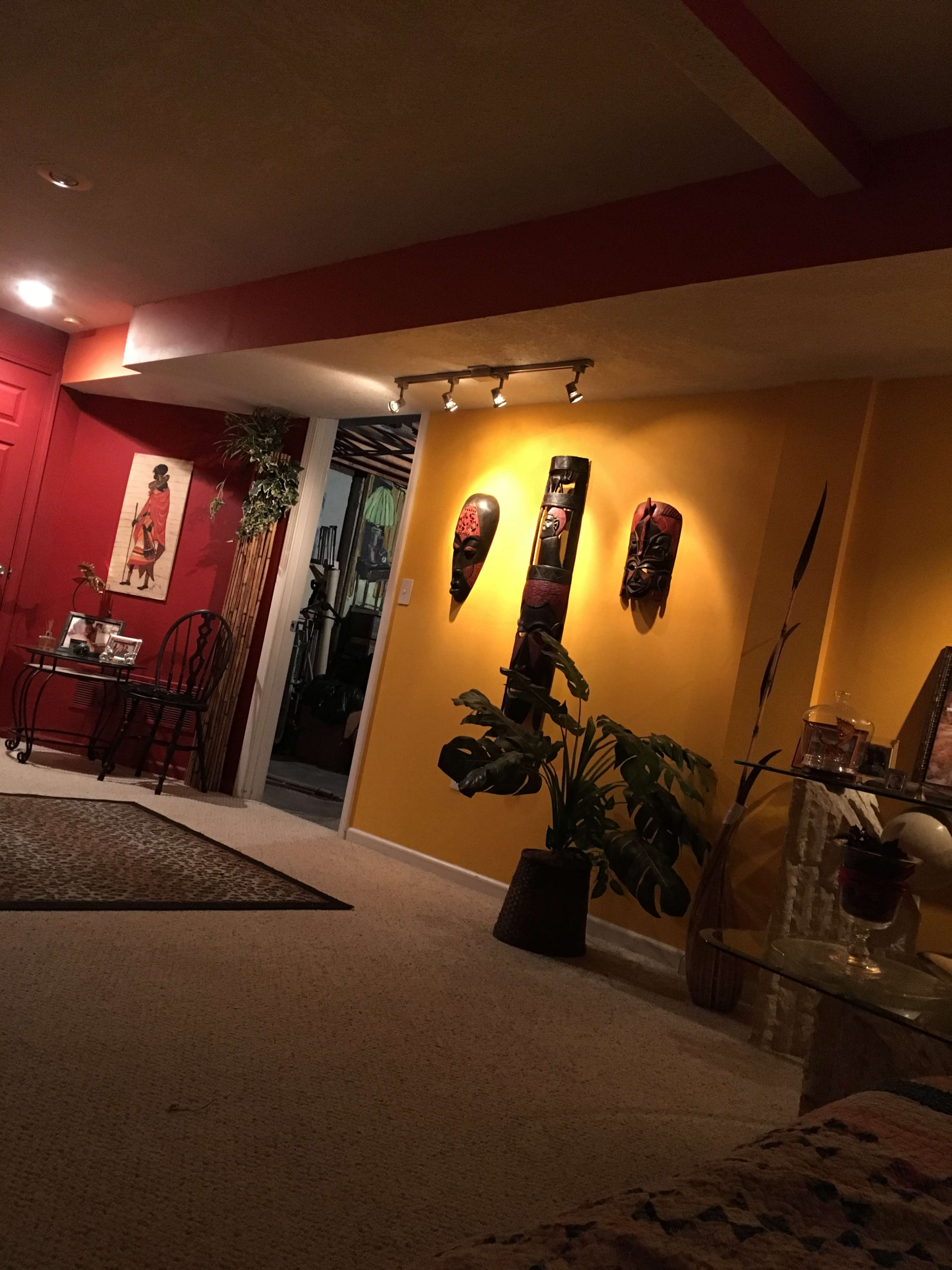 The image shows a warmly lit living space with a yellow wall adorned with three decorative masks, a potted plant in the foreground, and a red wall with a door in the background.