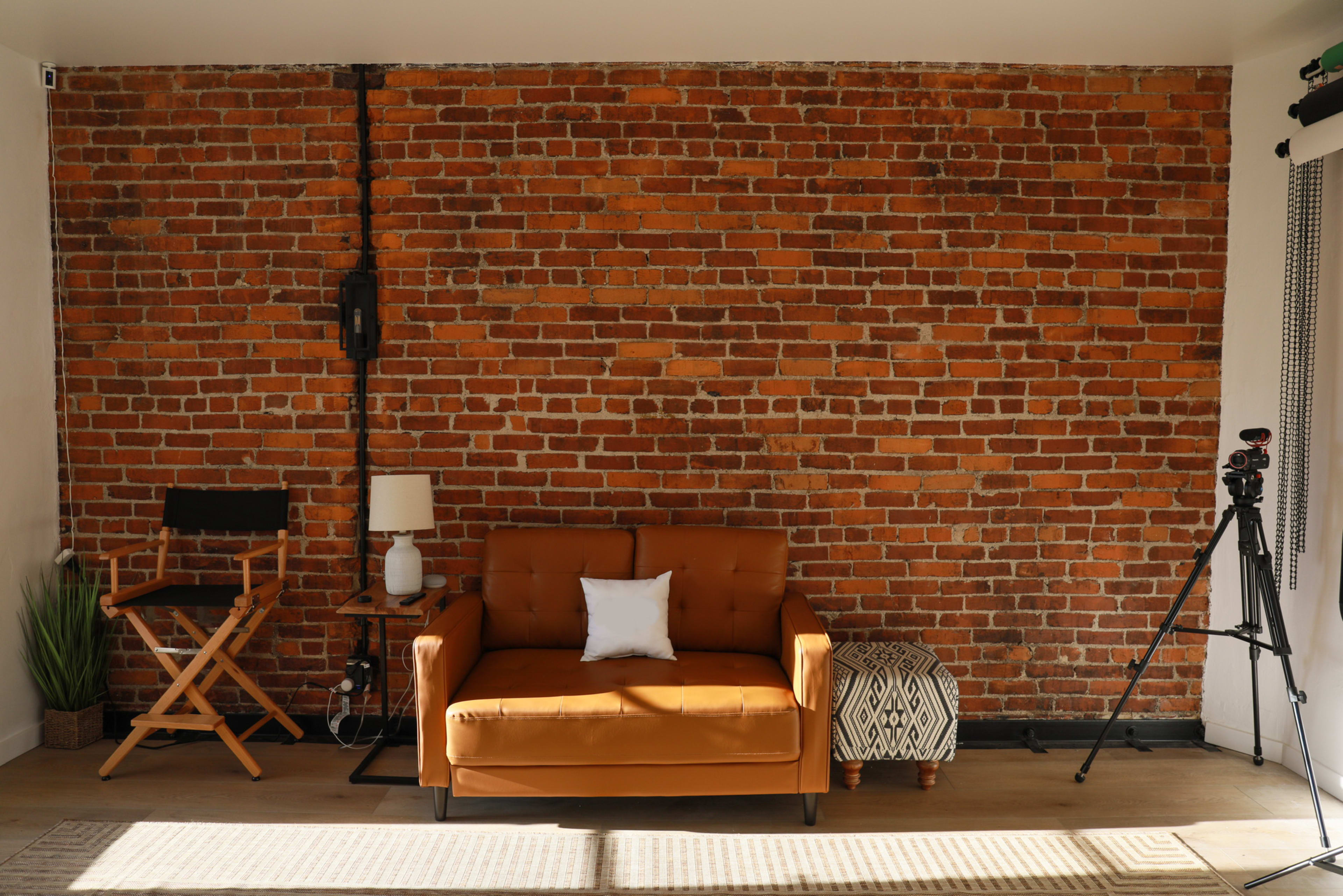 A modern interior featuring a brown leather sofa, a small patterned ottoman, a table lamp, a director's chair, a potted plant, and a tripod against a brick wall.
