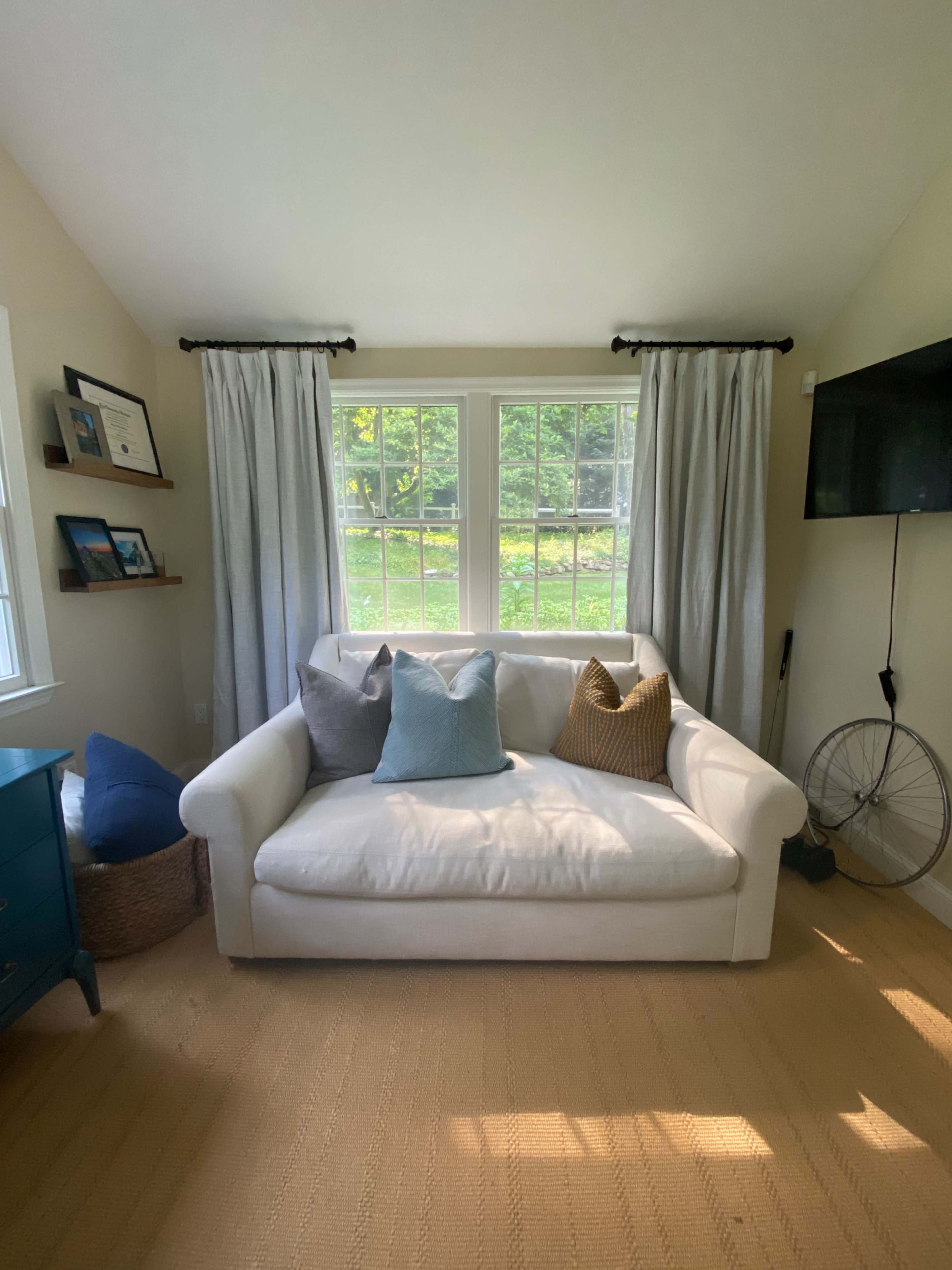 A white couch with colorful pillows sits in front of a large window, with light-colored curtains framing the view of a green outdoor area.