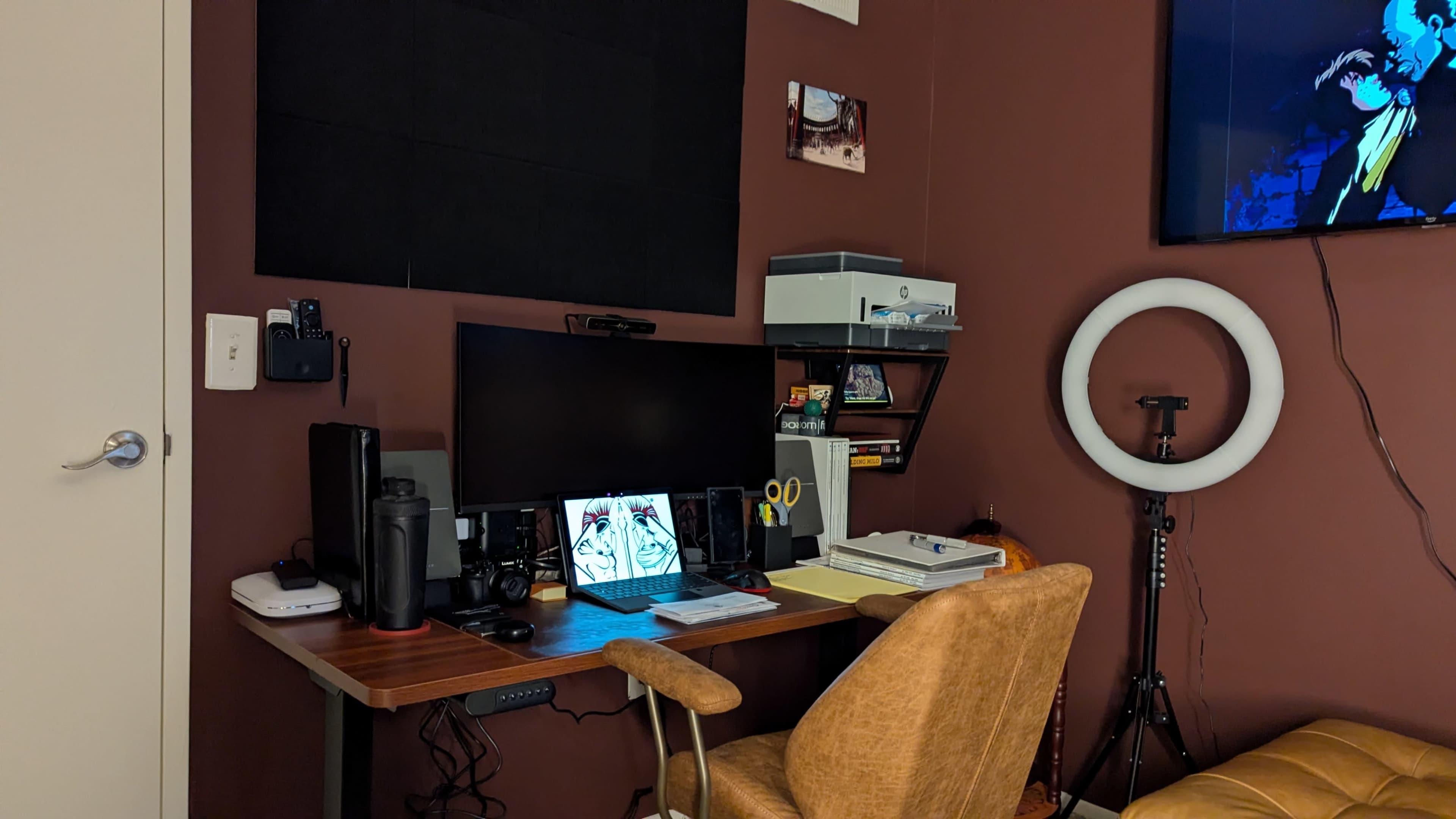 The image shows a home office setup featuring a wooden desk with a monitor, a laptop, various office supplies, a chair, and a ring light on a stand against a brown wall.