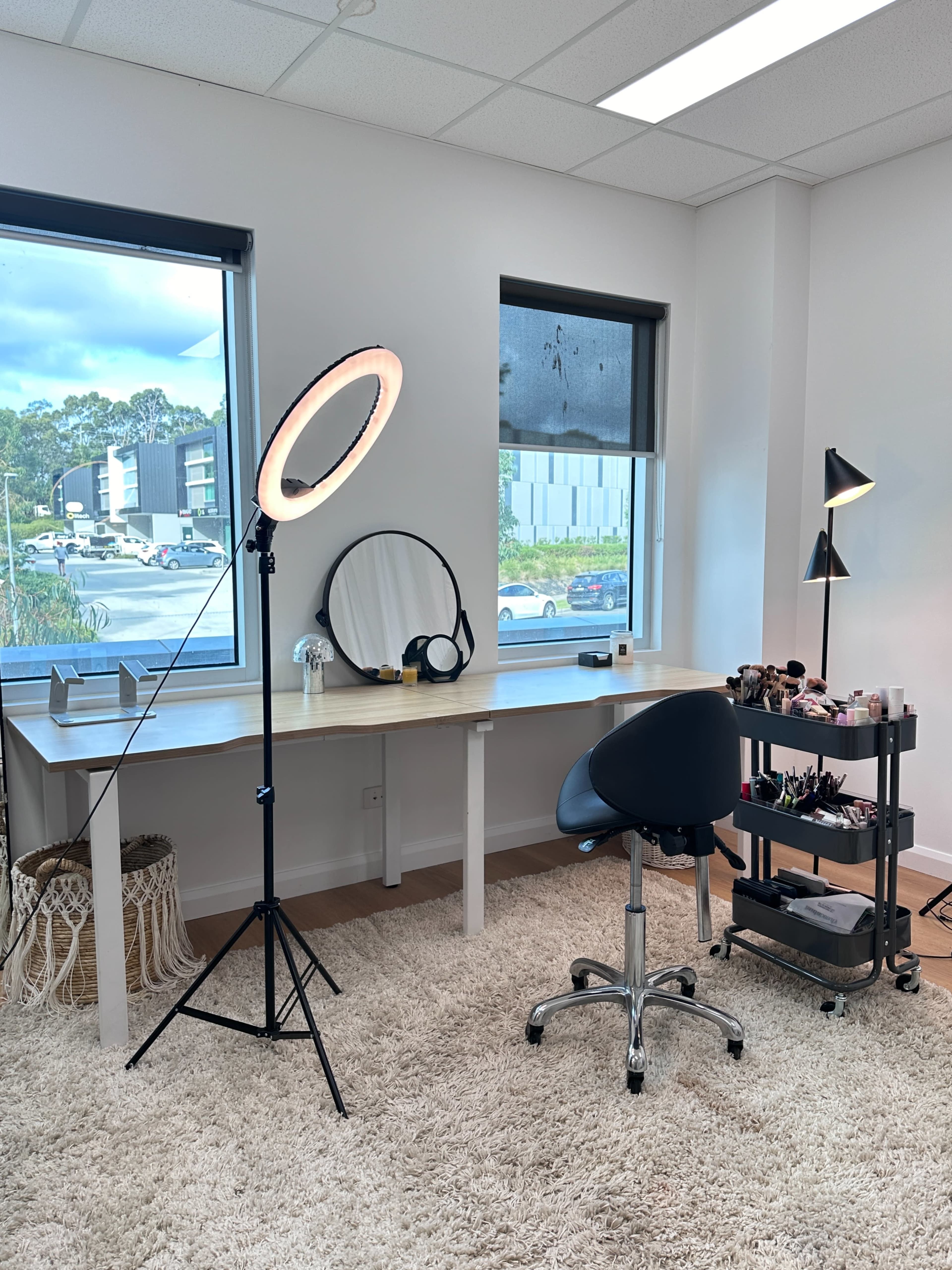 The image depicts a bright, organized makeup studio featuring a ring light, a mirror on a wooden table, a rolling cart with makeup products, and a chair on a soft rug.