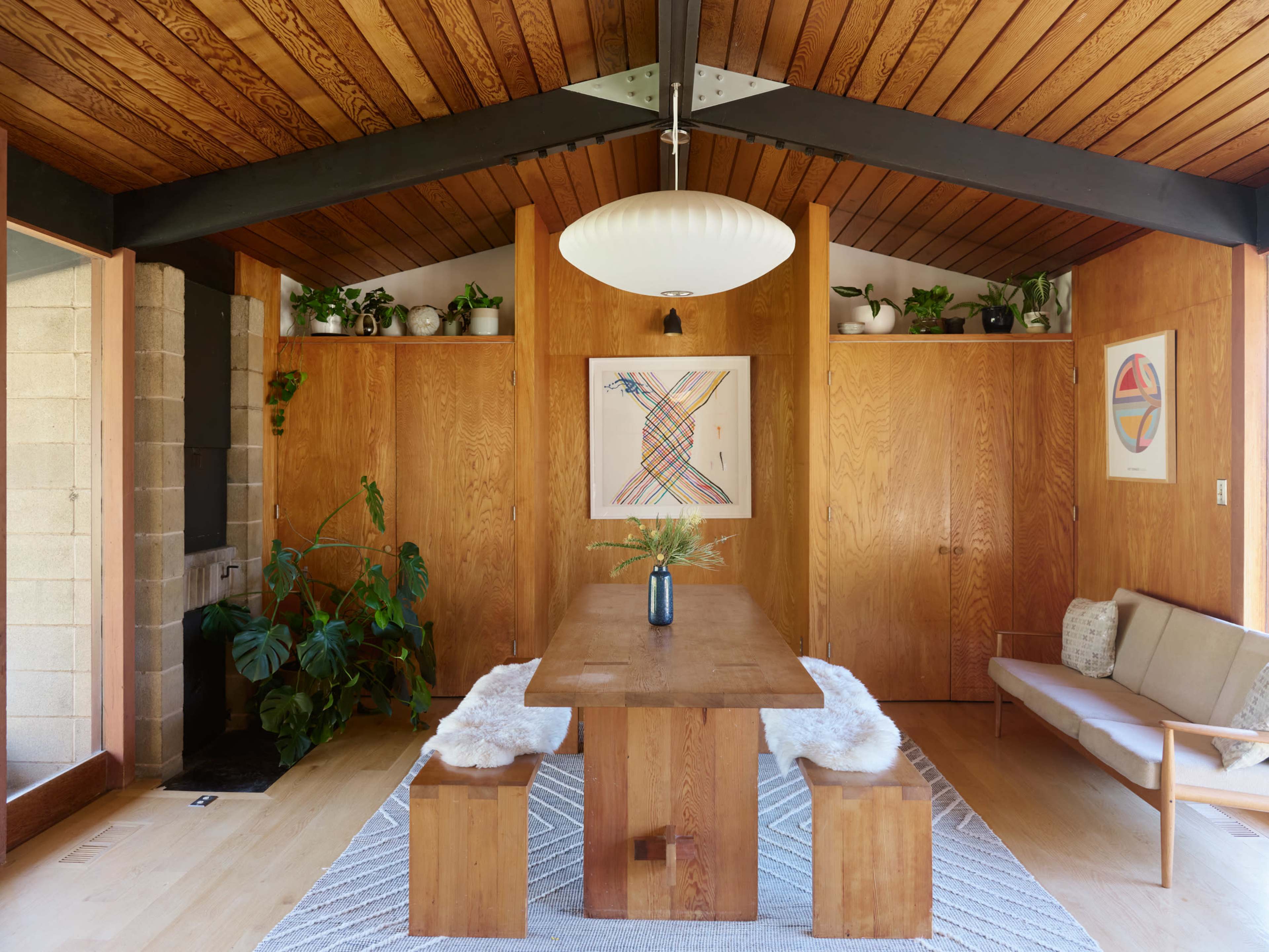 A wooden dining table with benches is centered in a room featuring wood-paneled walls and a high ceiling with exposed beams, adorned with plants and artwork.