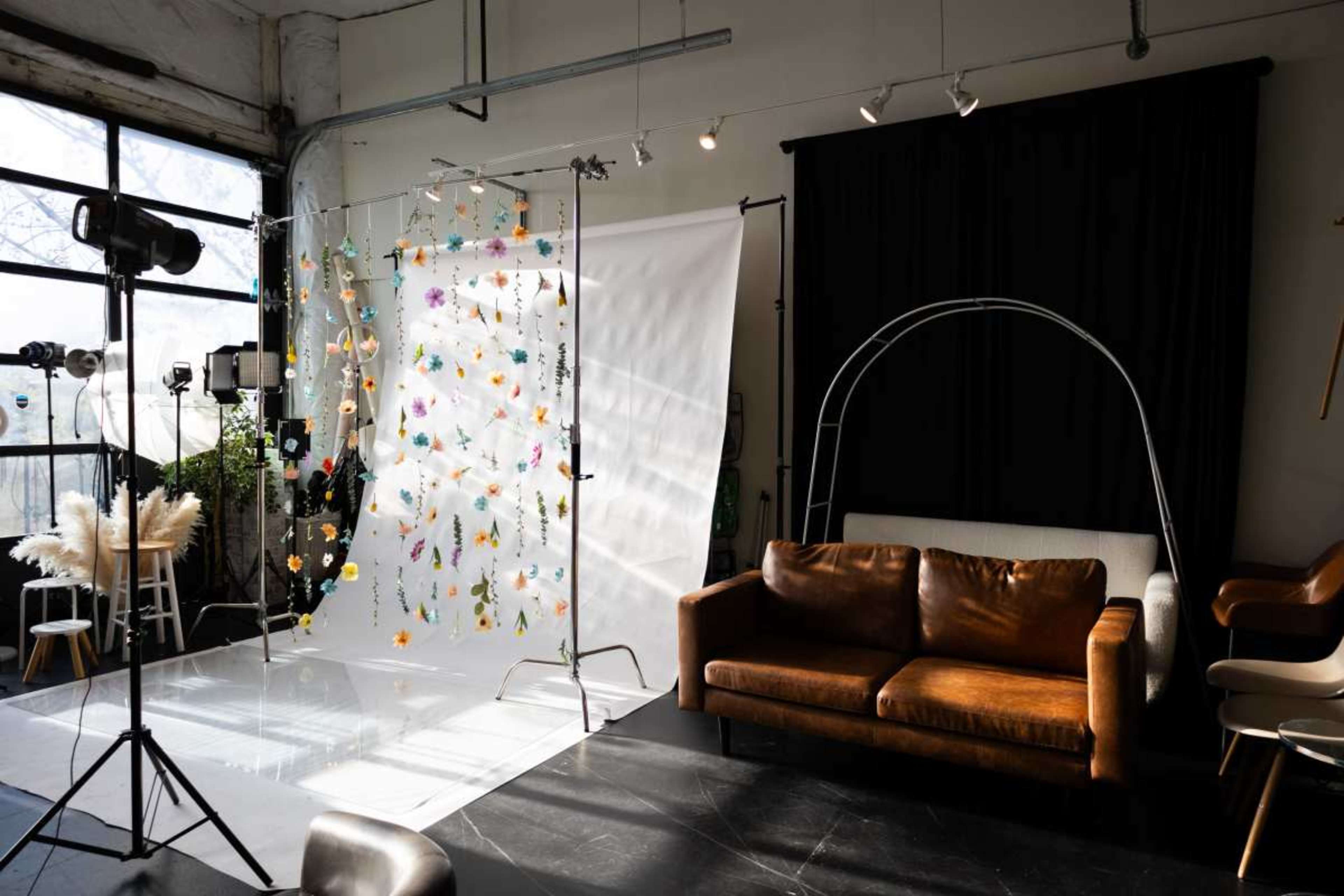The image shows a photography studio featuring a colorful backdrop, a large white screen, a brown leather sofa, and various lighting equipment.