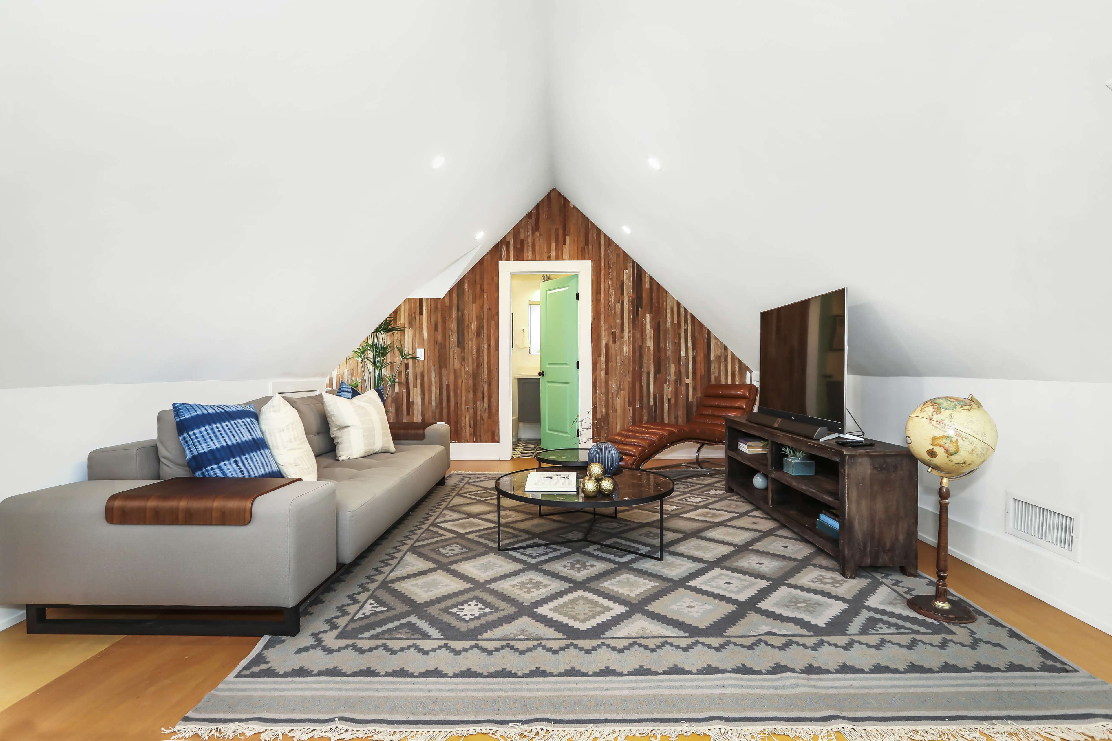 The image shows a cozy attic living room featuring a gray sectional sofa, a television on a wooden stand, and a geometric area rug beneath a coffee table.