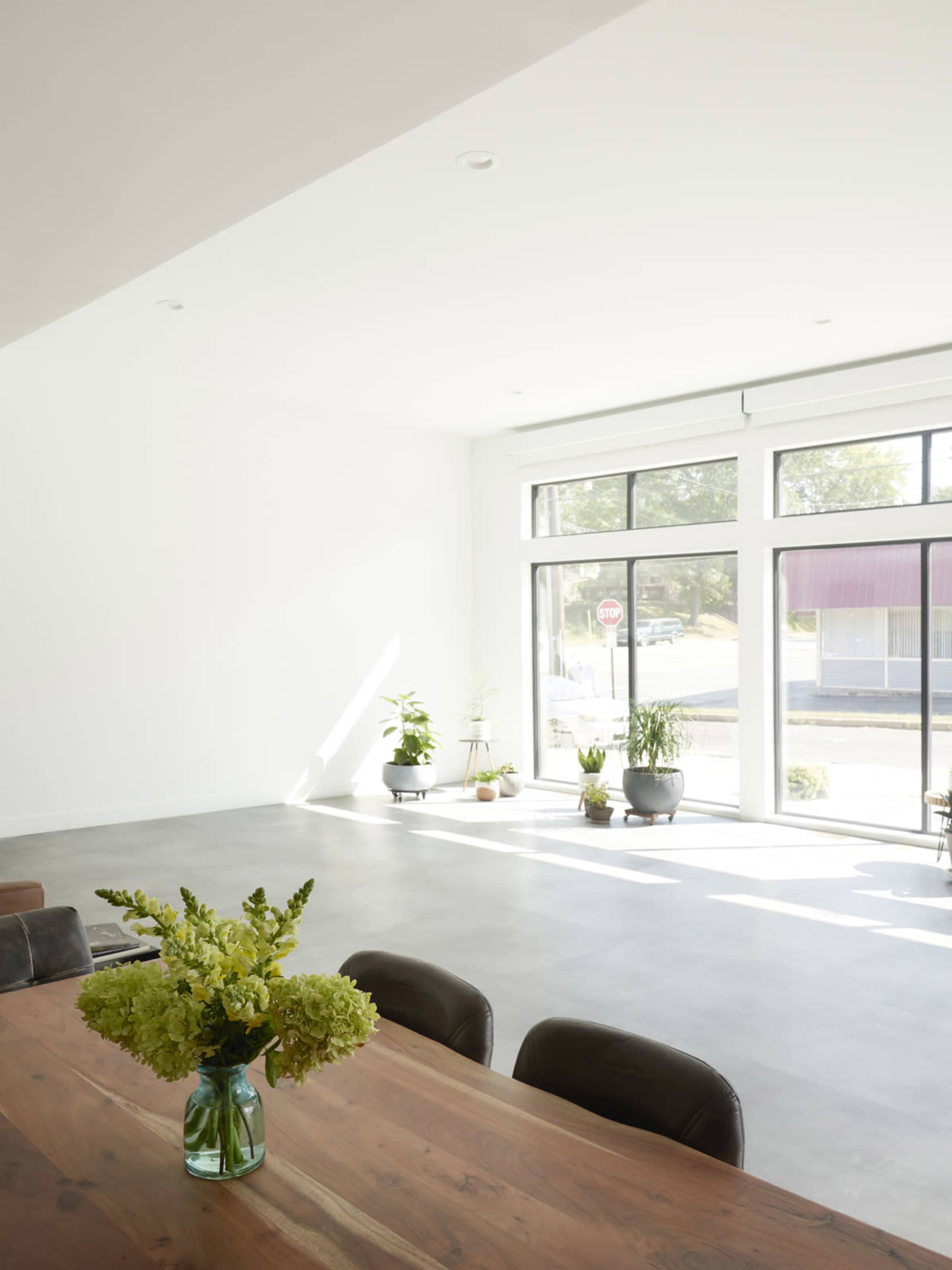 A spacious room with large windows showcasing plants and a wooden dining table with a flower vase in the foreground.