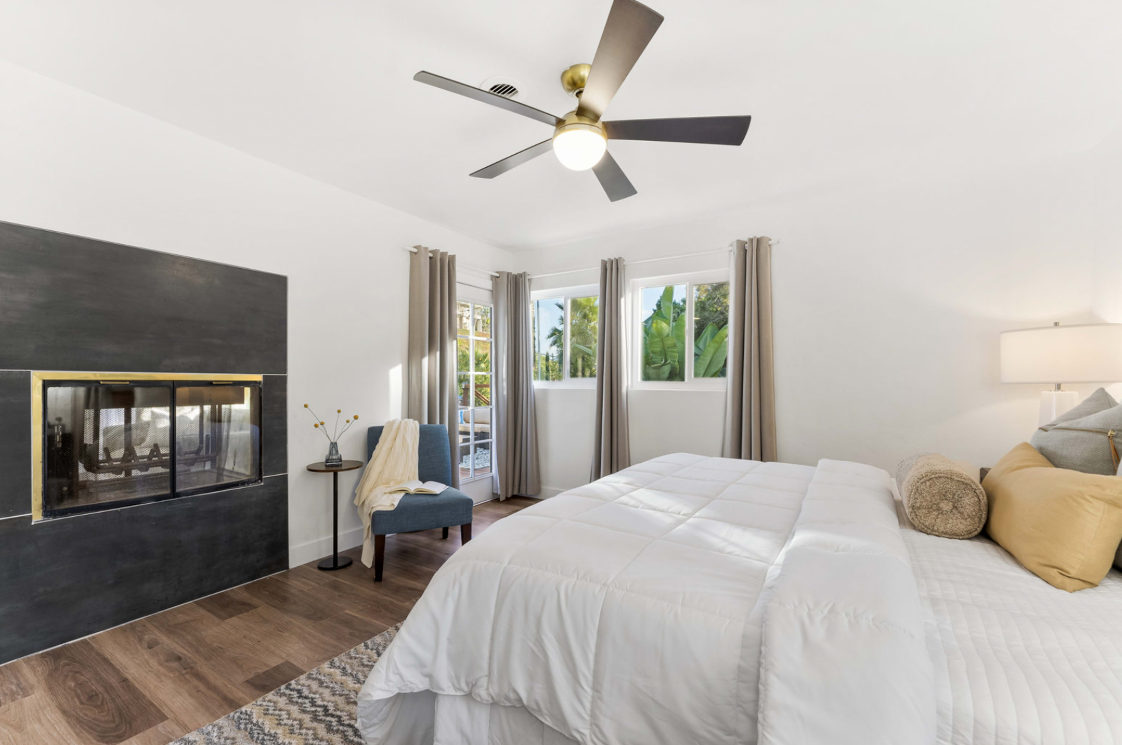 The image shows a modern bedroom featuring a bed with white bedding, a fireplace with a black surround, a ceiling fan, and large windows with curtains.