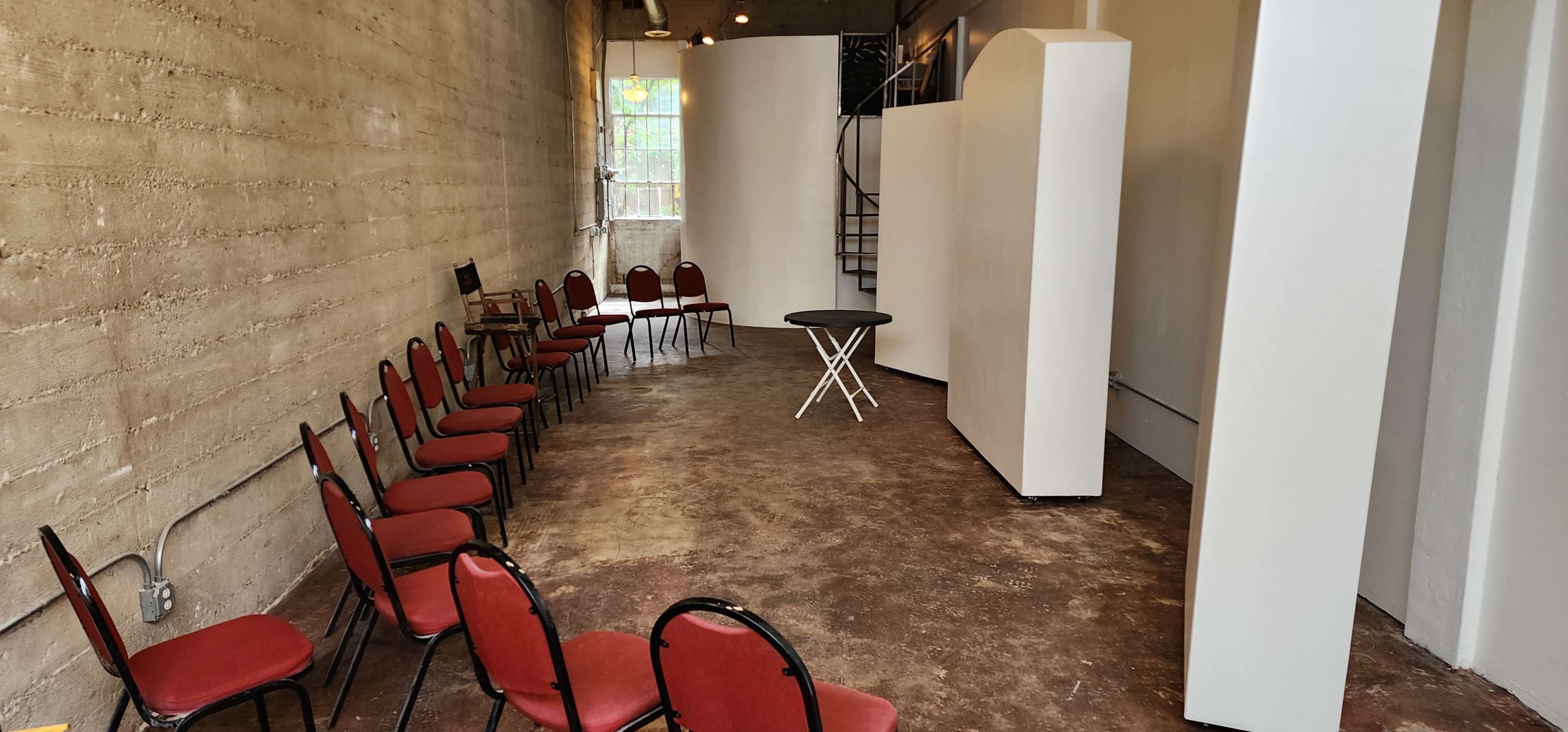 An empty room with a row of red chairs arranged around a small black table, alongside white wall partitions and a staircase in the background.