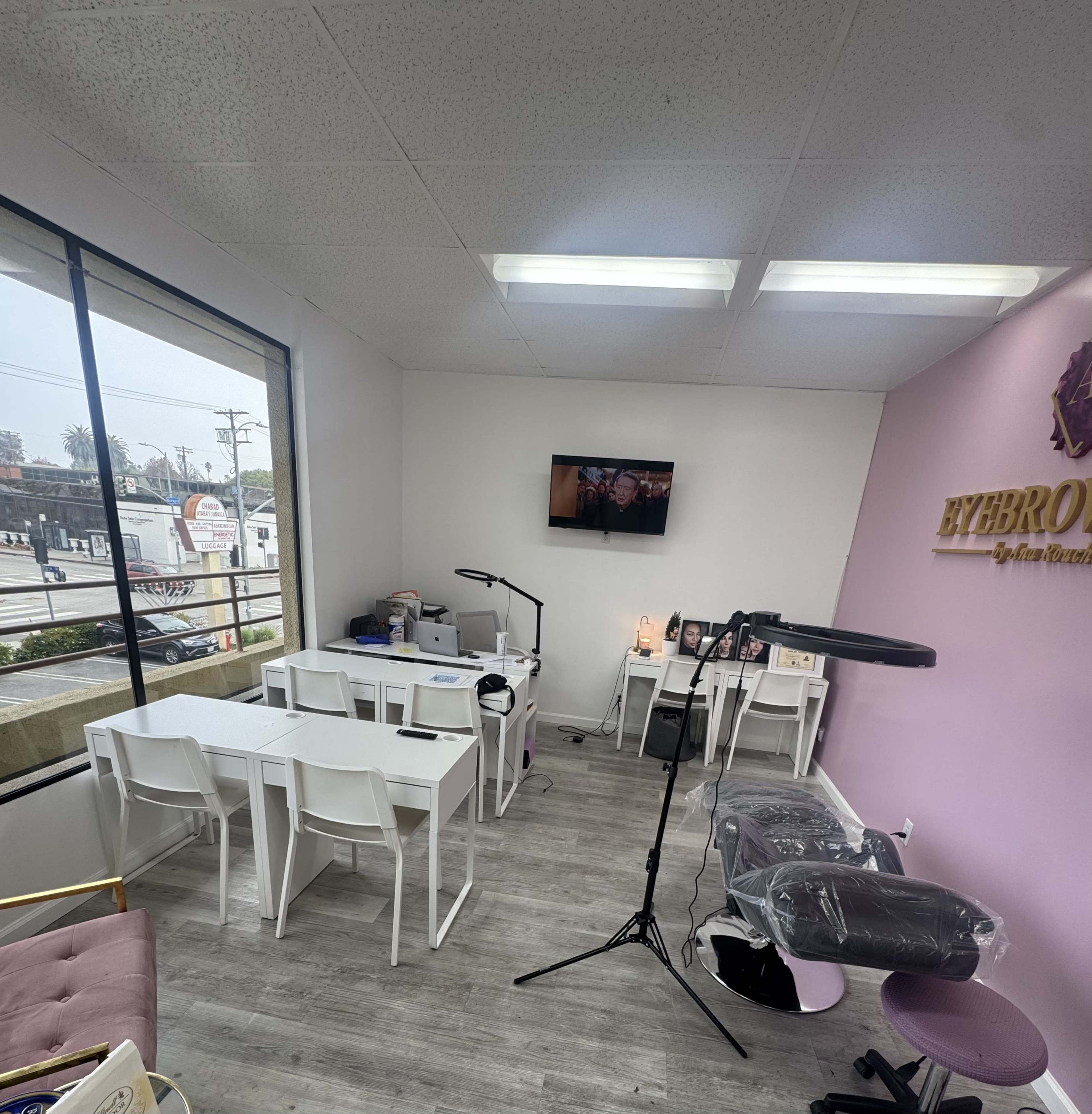 The image shows a small, modern beauty studio with several white tables, an area for clients, and a wall-mounted television.