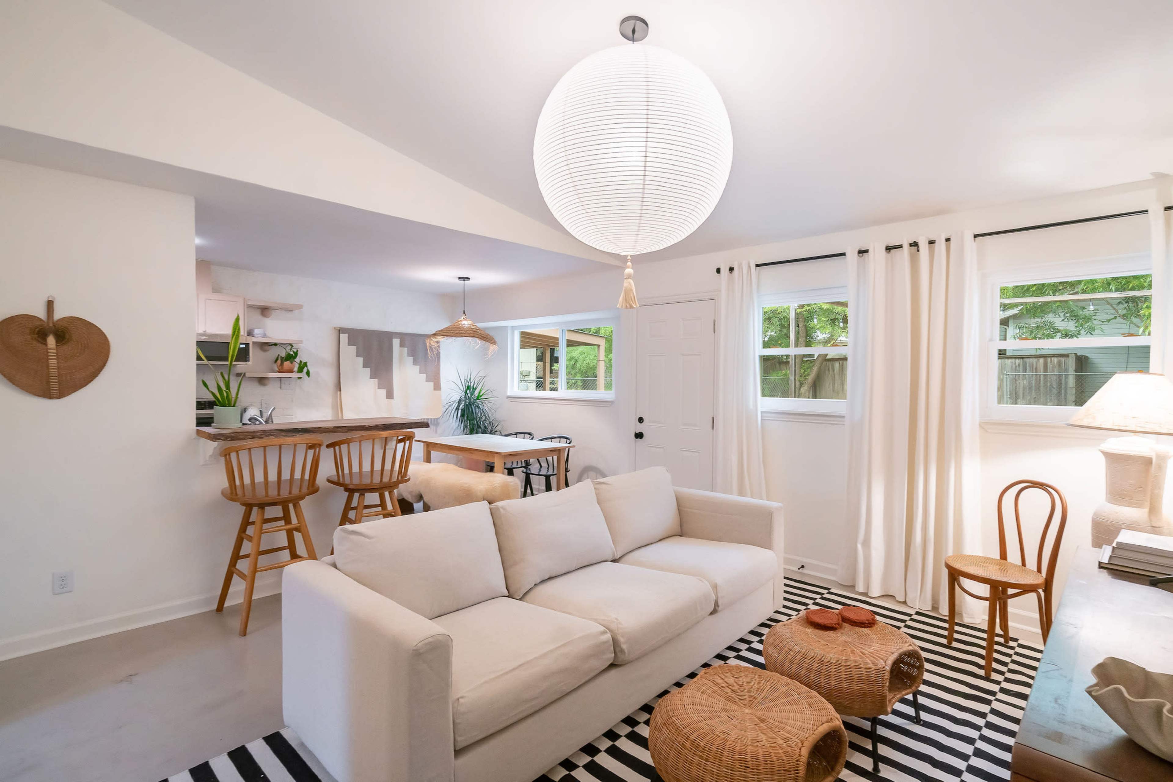 A modern living area with a white sofa, a black and white striped rug, a dining bar with wooden stools, and large windows allowing natural light.