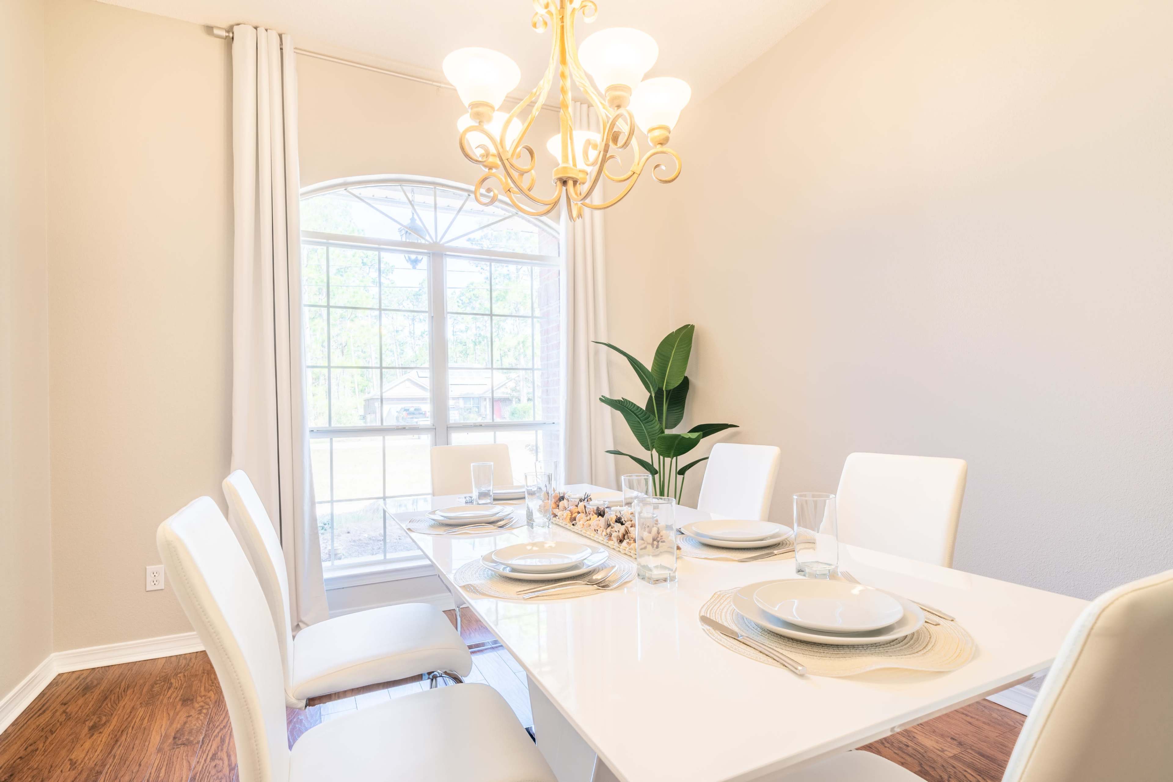 A modern dining room features a white table set for six, with a chandelier above and a large window providing natural light.