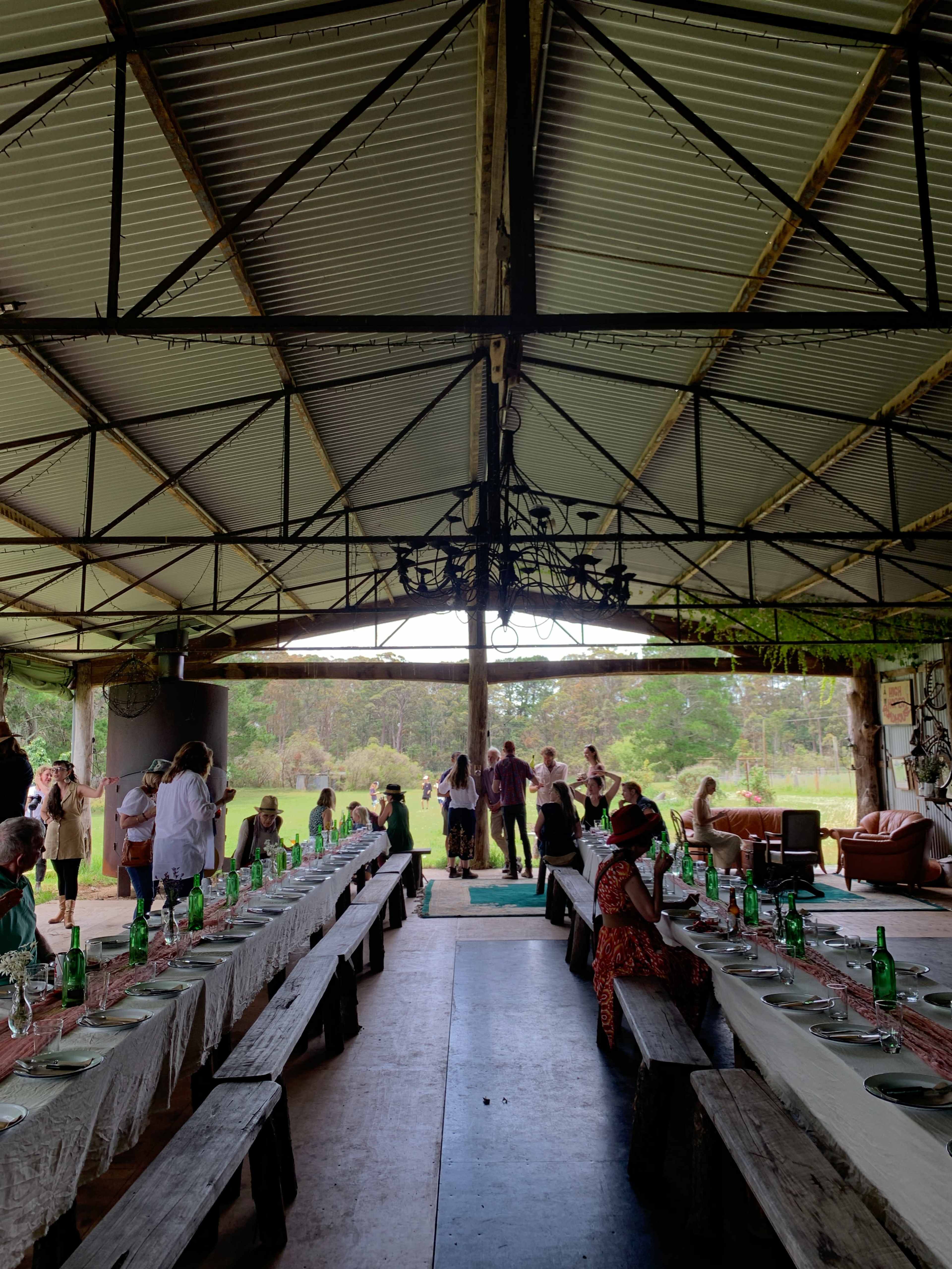 A spacious dining area features long tables set with green bottles and tablecloths, while guests mingle near an open entrance overlooking a natural landscape.
