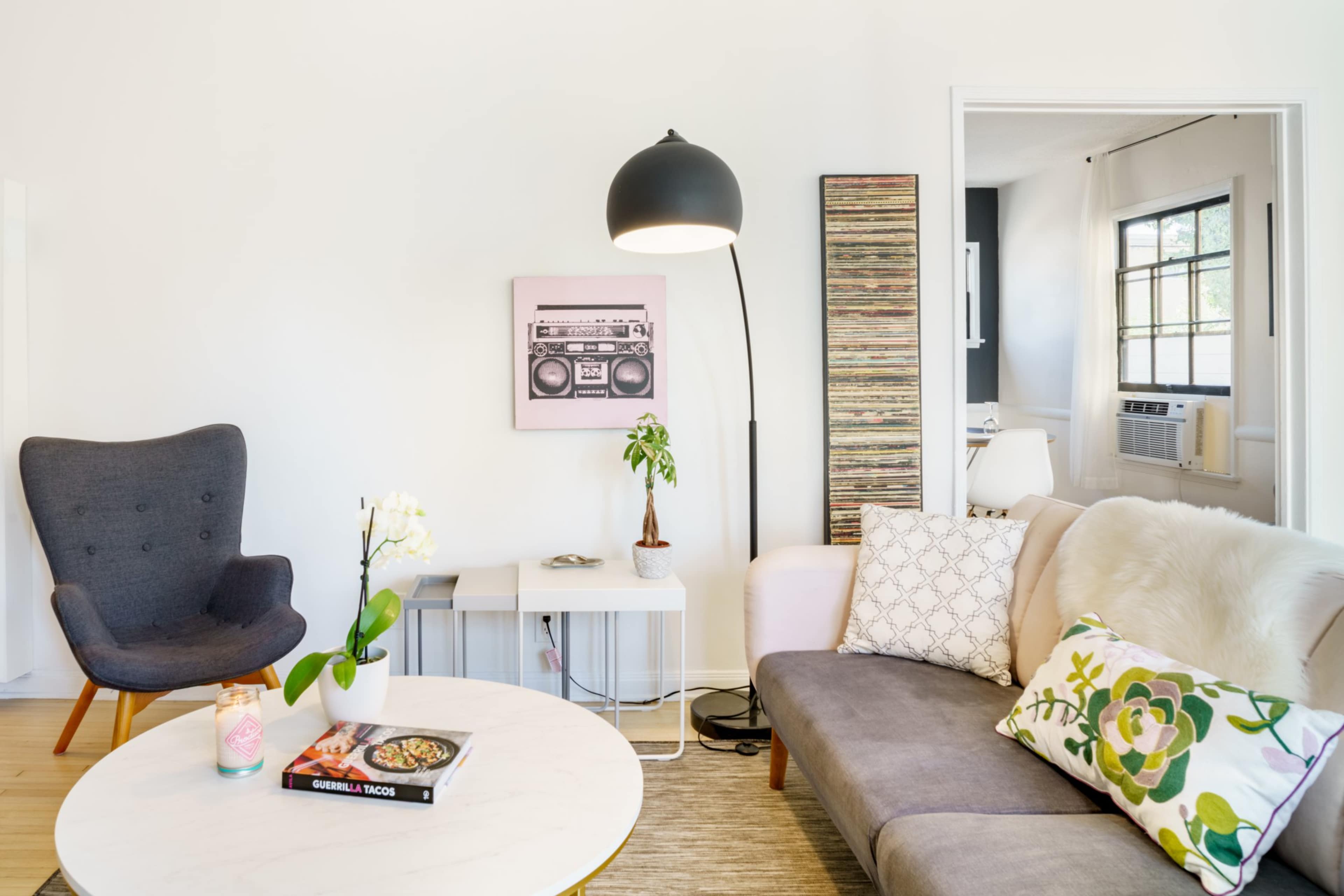 The image shows a modern living room with a gray sofa, a comfortable armchair, a round coffee table, and a floor lamp, featuring decorative plants and artwork on the walls.