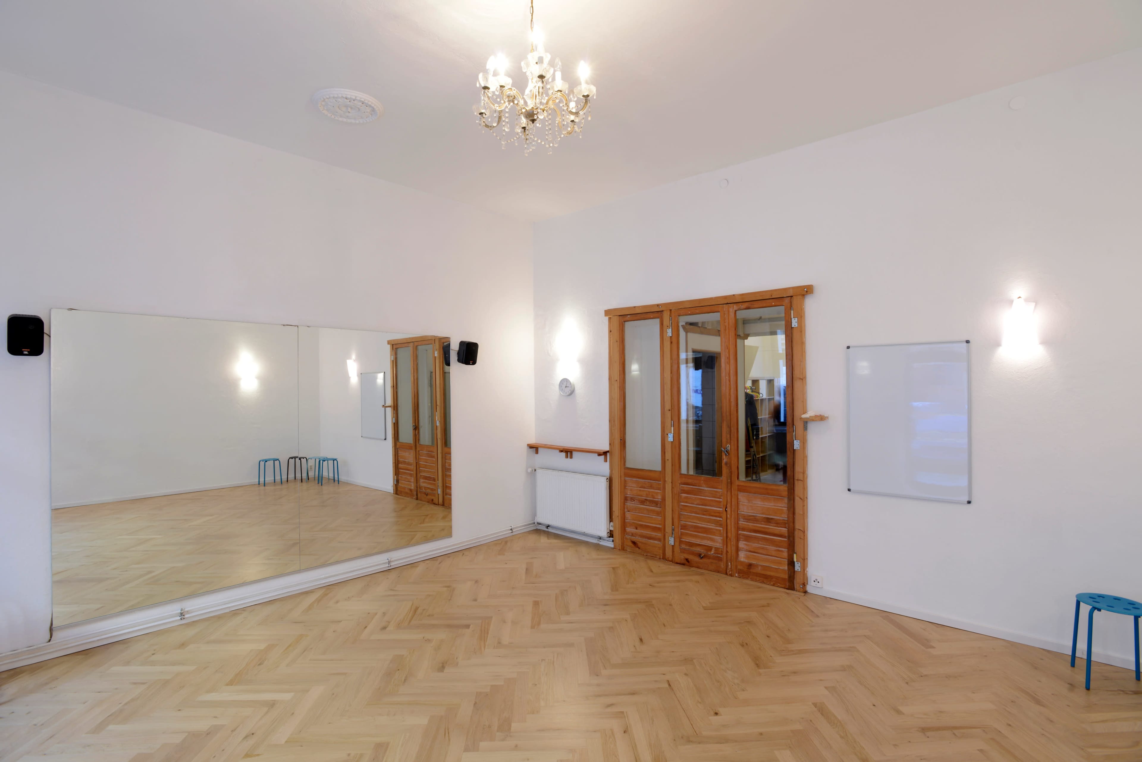 A spacious room with wooden flooring, a large mirror, a chandelier, double wooden doors, and a whiteboard on the wall.