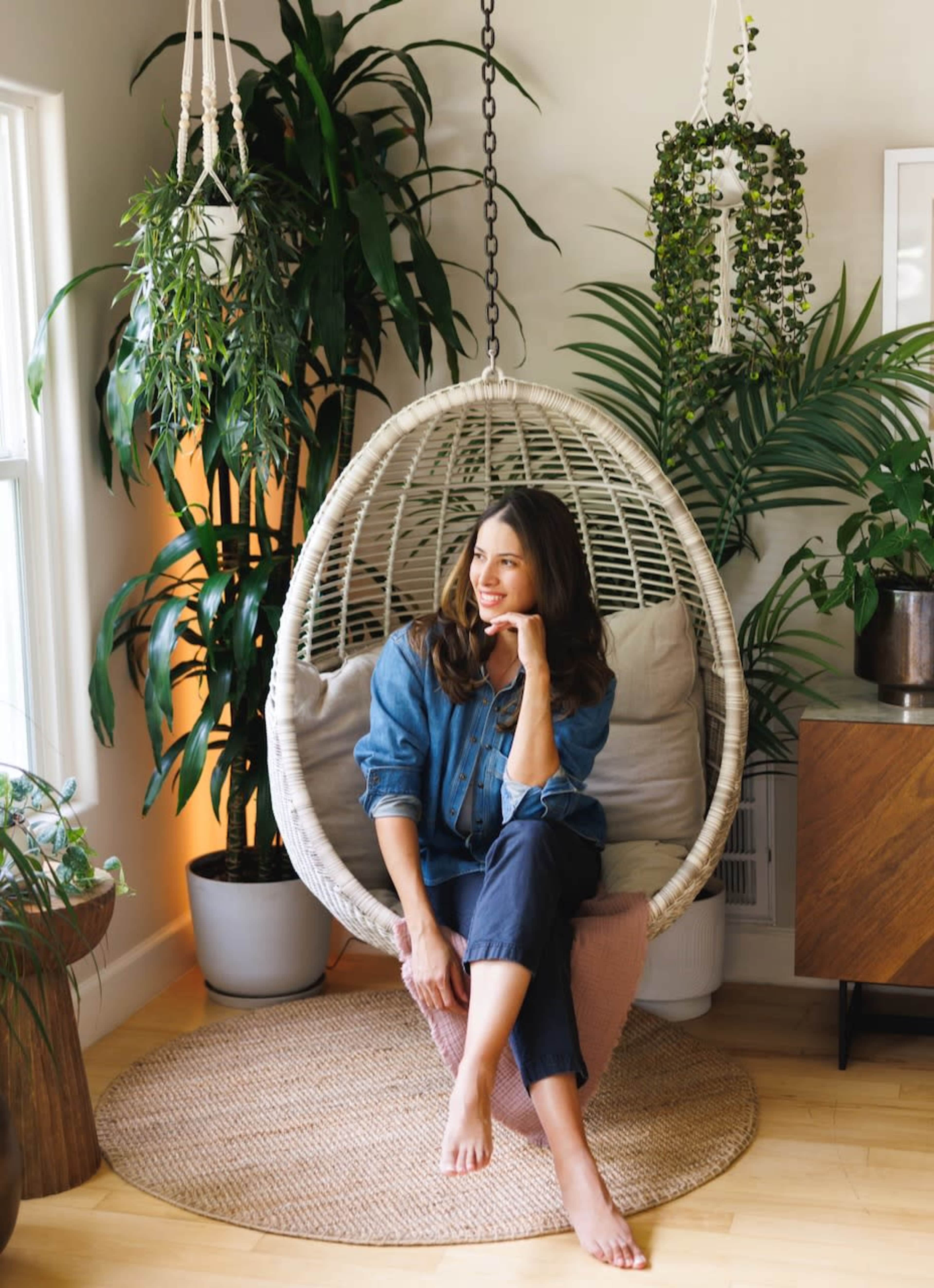 A woman sits in a hanging chair surrounded by large indoor plants.
