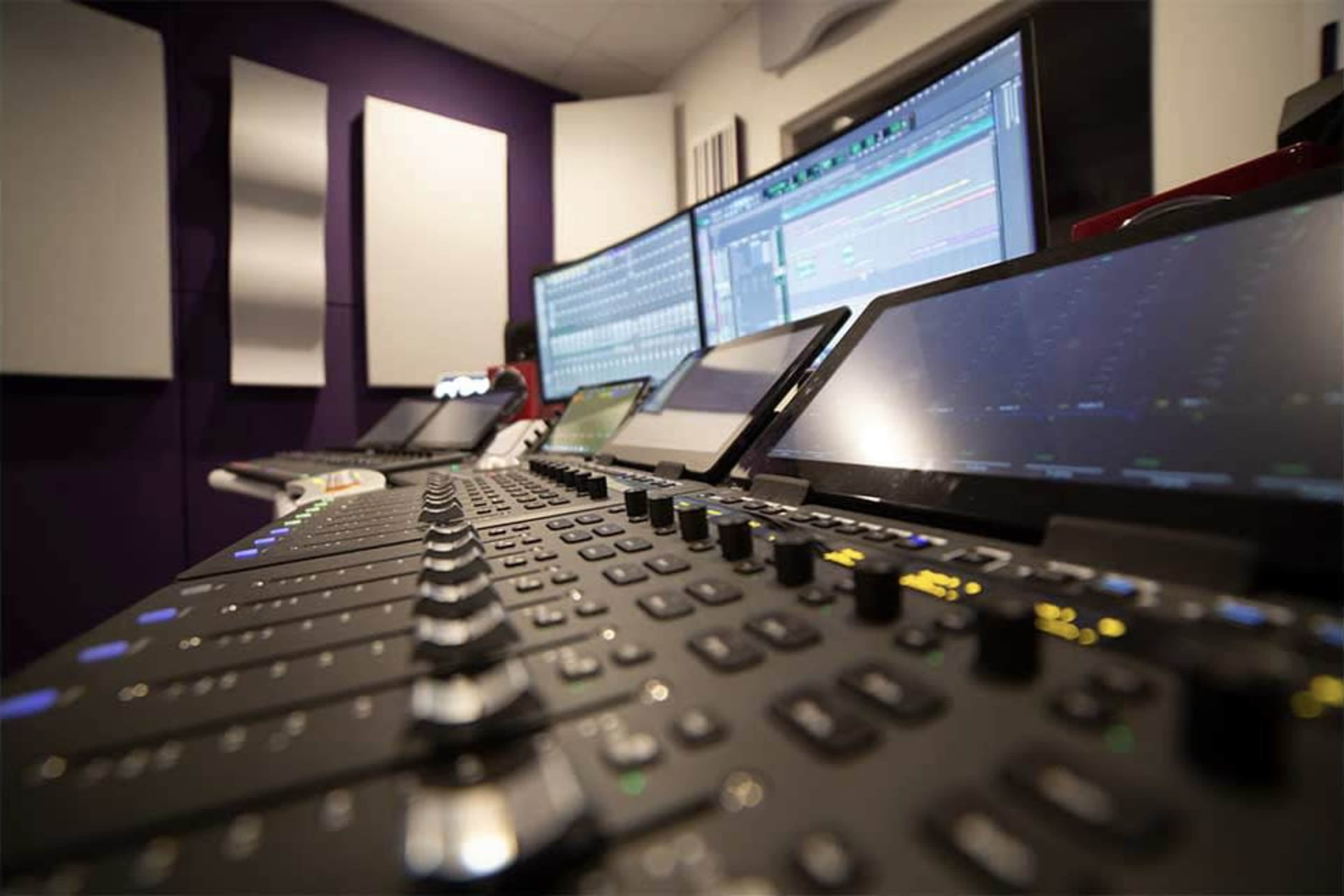 A modern music production studio with multiple computer screens displaying audio editing software and a control panel featuring various knobs and sliders.