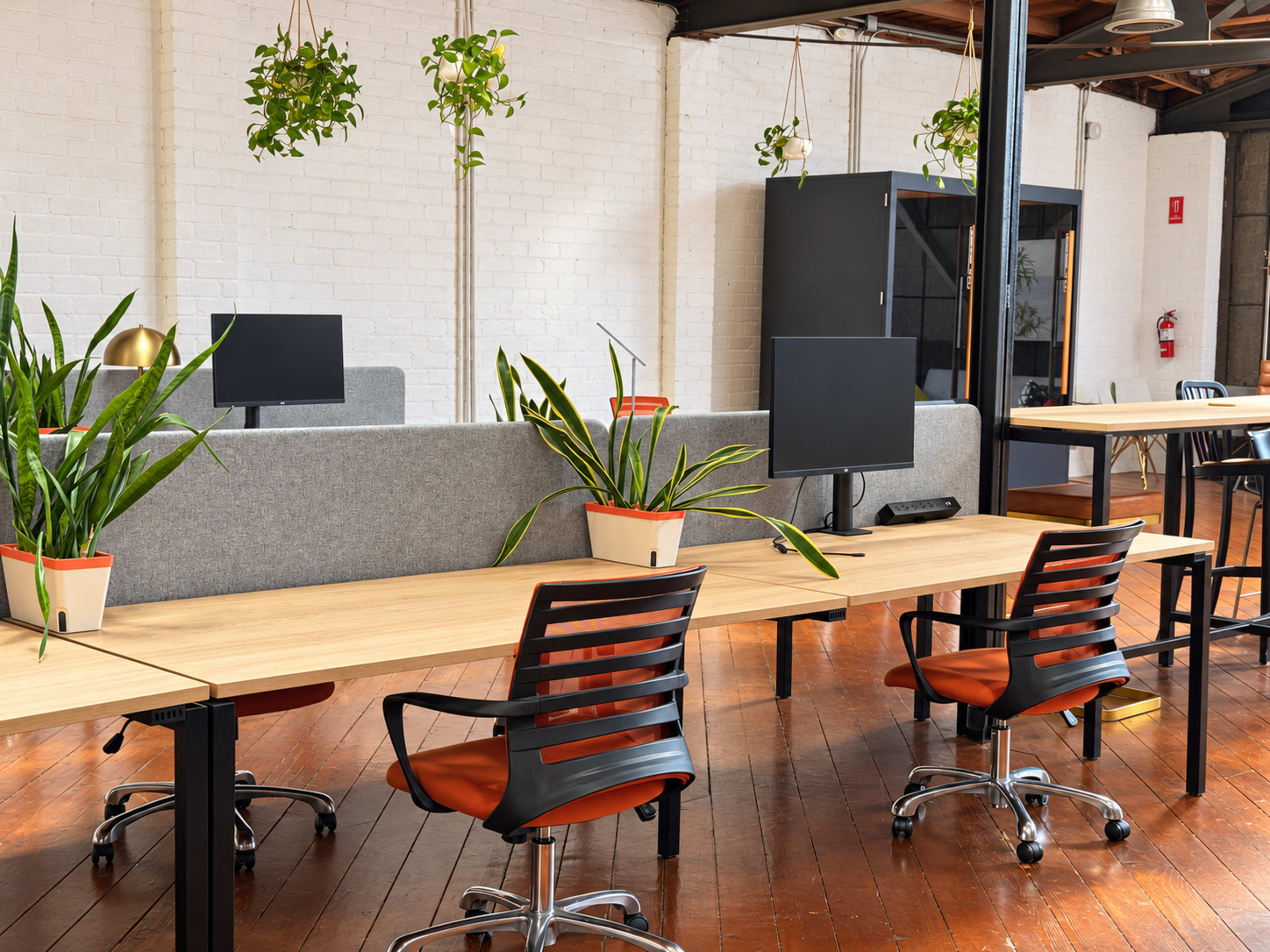 The image shows a modern office space with several desks equipped with plants, black monitors, and ergonomic chairs arranged in a spacious area with wooden flooring and white brick walls.