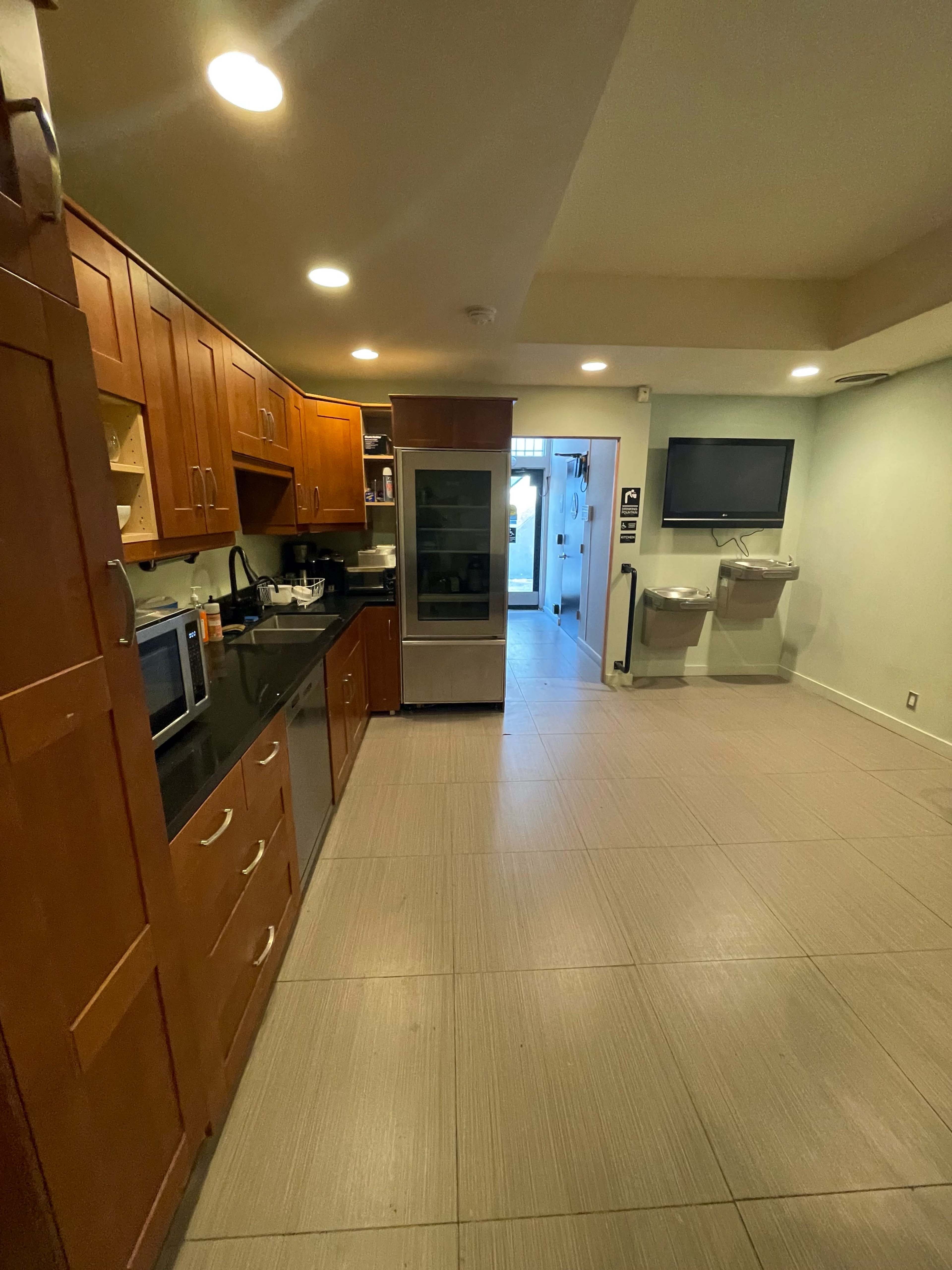 A modern kitchen area with wooden cabinets, a black countertop, a refrigerator, a microwave, and a door leading to an outdoor space, alongside two water fountains.