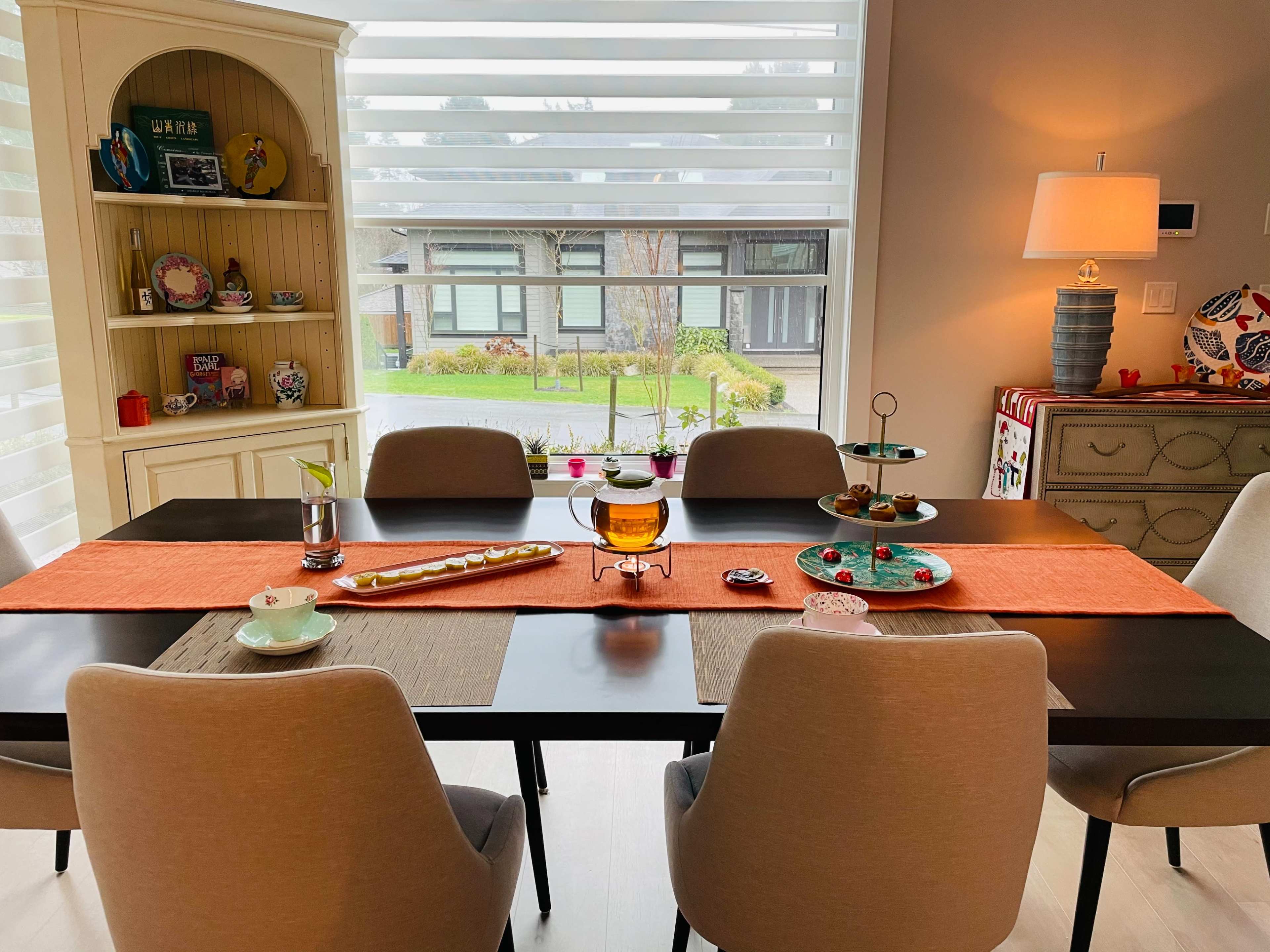 A dining table is set with various items, including a centerpiece and table runners, surrounded by chairs in a brightly lit room with large windows.