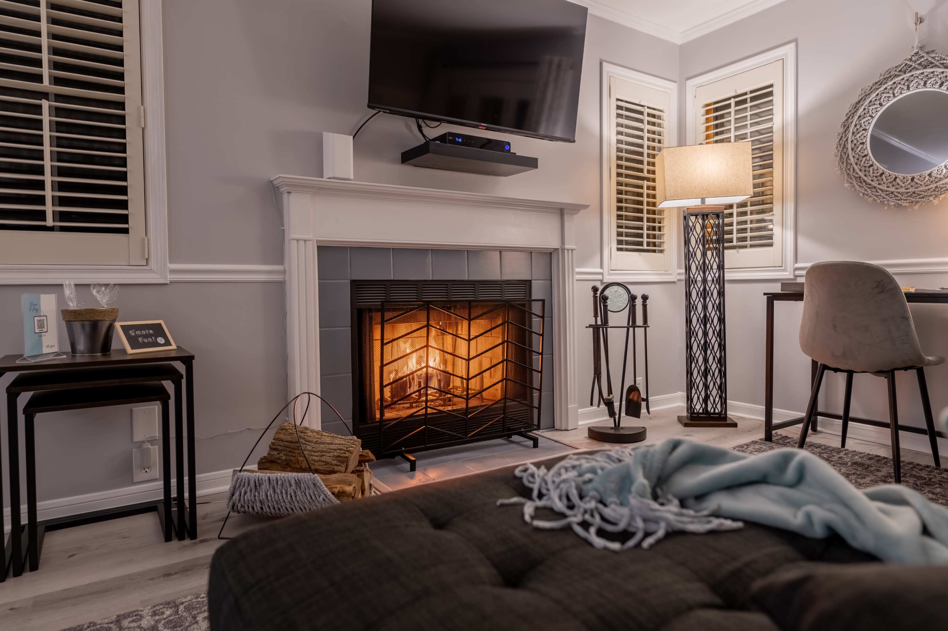 A cozy living space featuring a fireplace with a warm glow, a modern television mounted above it, a floor lamp, and a desk with a chair.