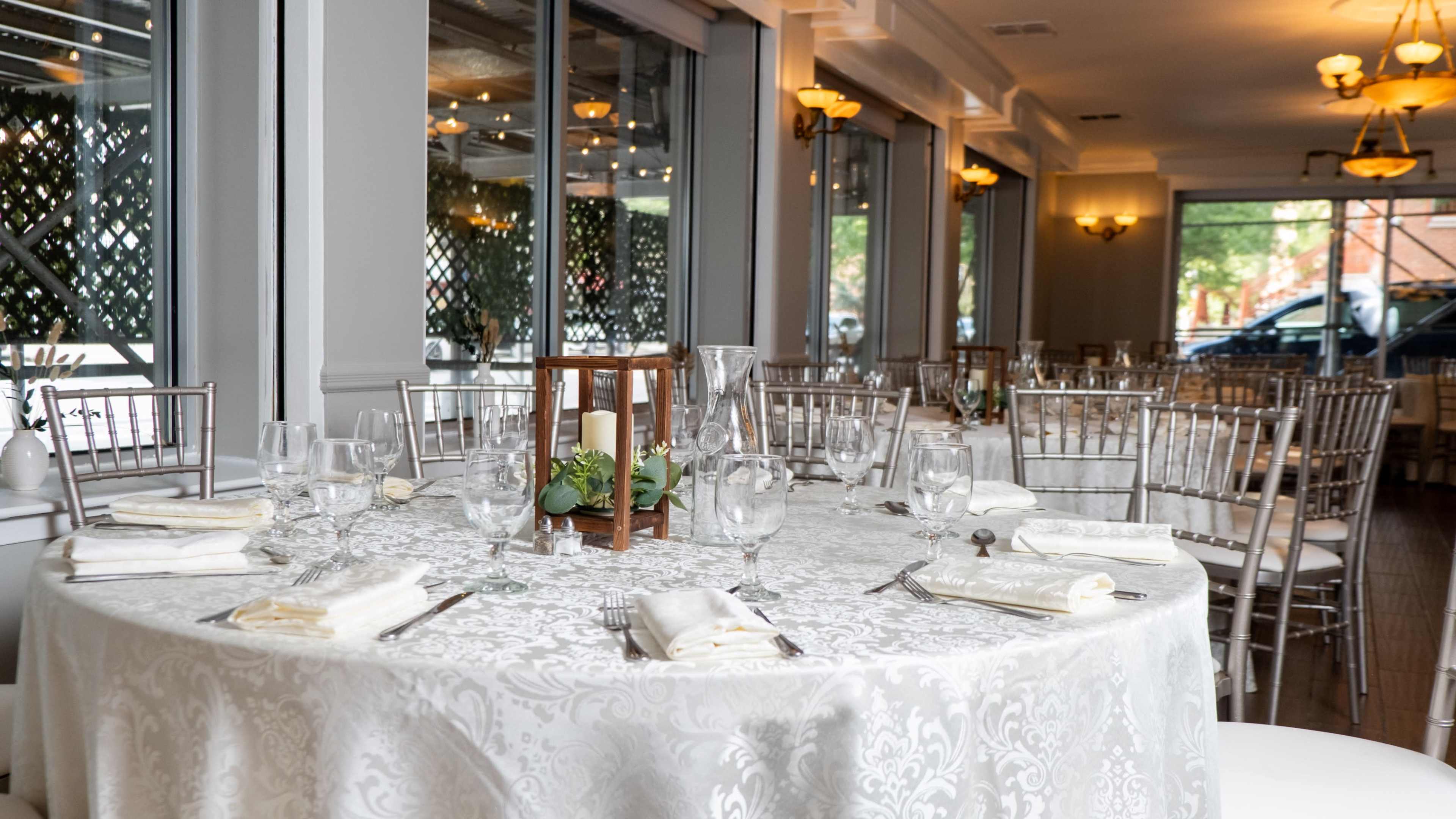 The image shows a dining setup in a elegantly decorated restaurant with a round table covered in a white lace tablecloth and surrounded by silver chiavari chairs.