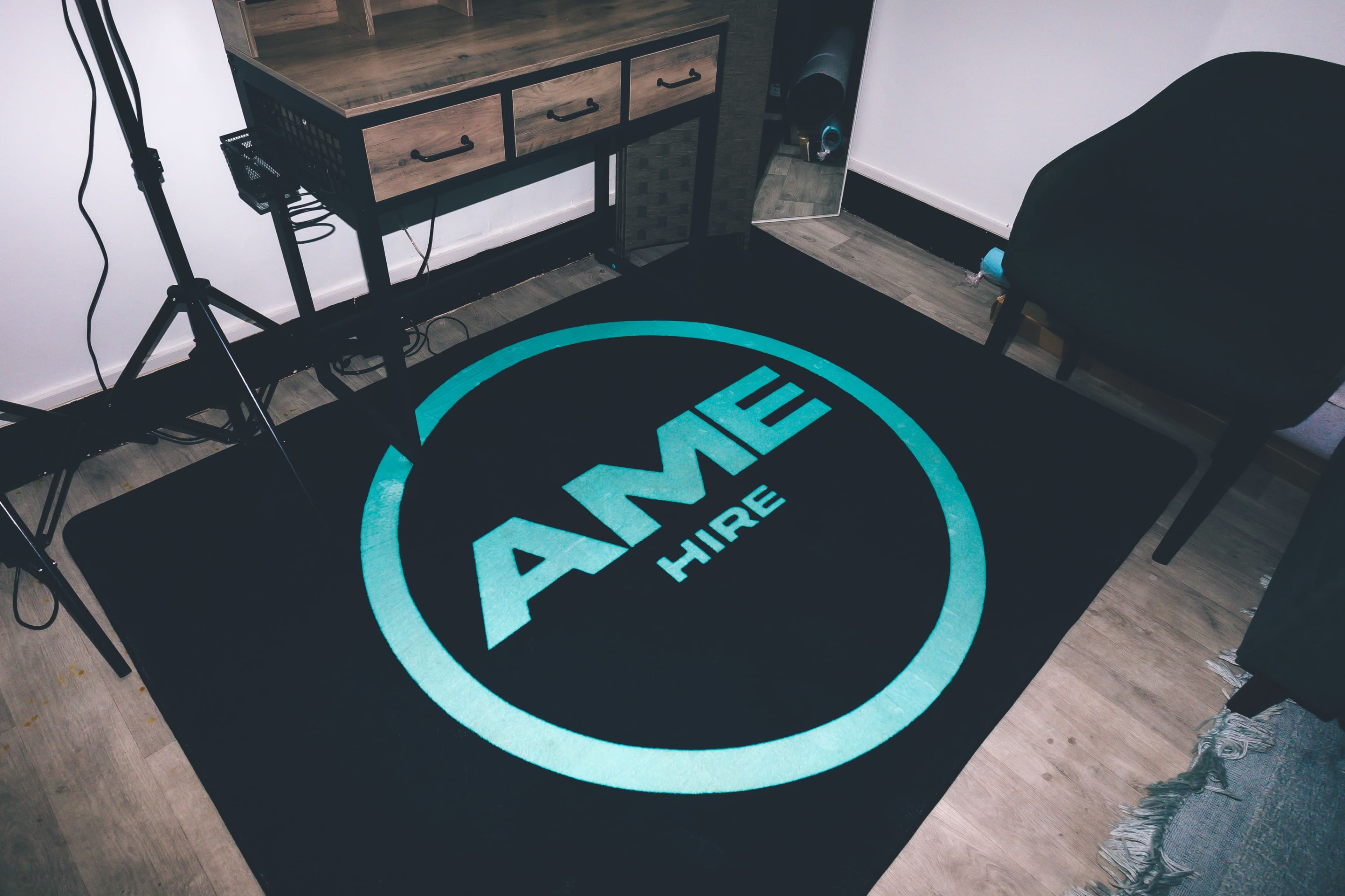 The image shows a black rug with a circular logo that reads "AME HIRE," situated on a wooden floor beside a desk and a chair.