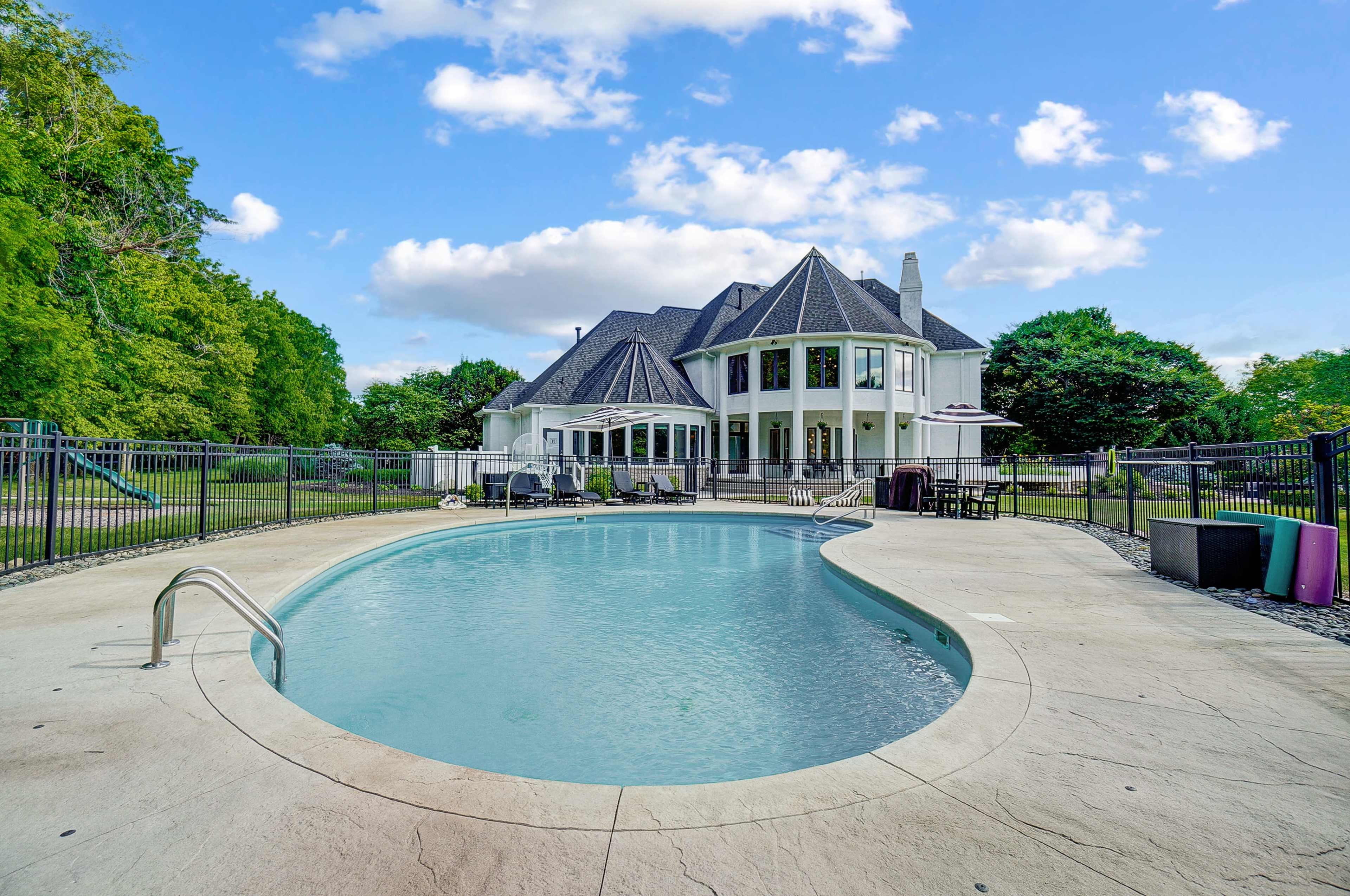 A large, modern house with a swimming pool in the foreground and a fenced backyard surrounded by trees.