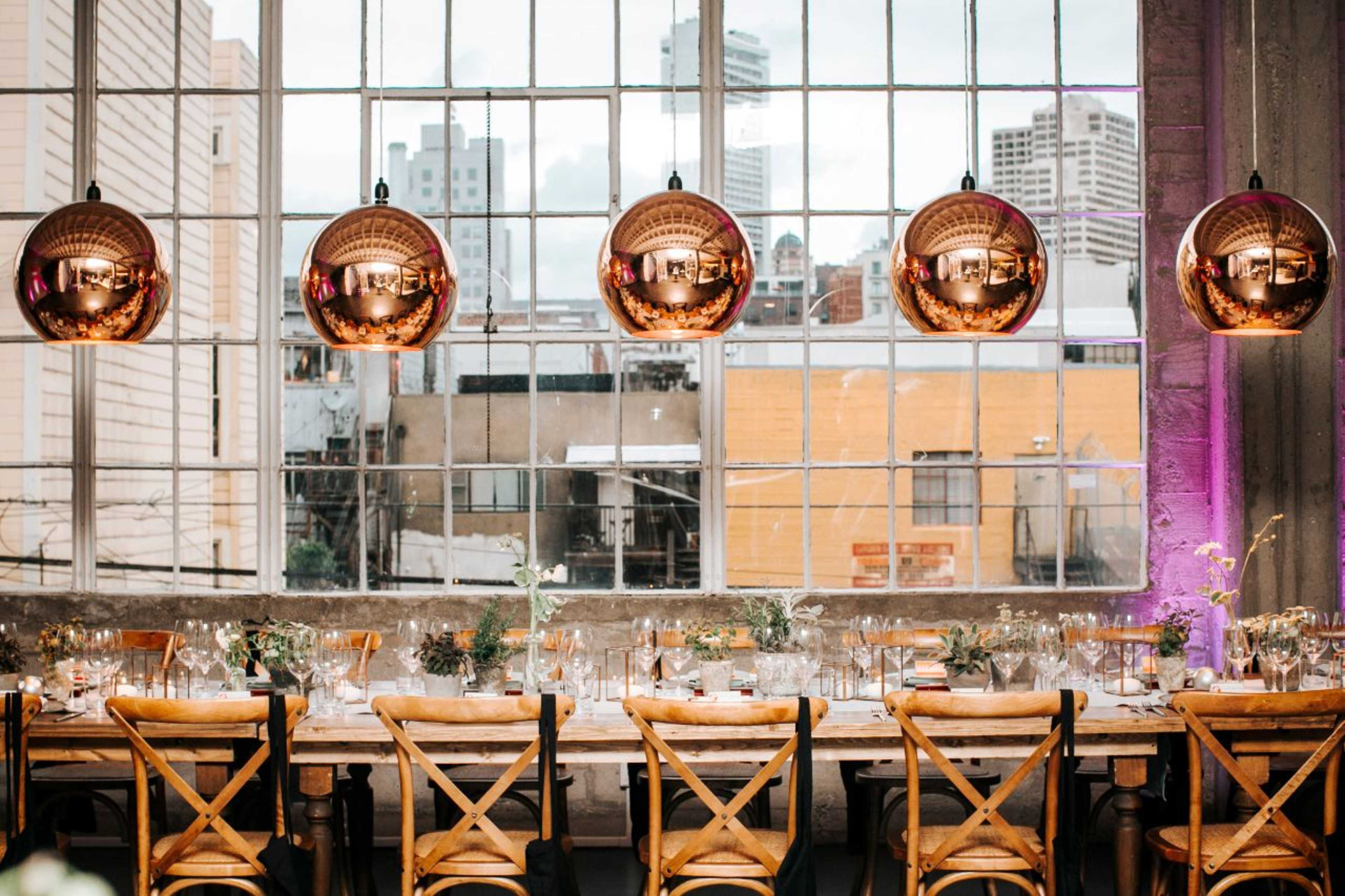 A long dining table is set with glasses and flowers, illuminated by large, round pendant lights hanging from the ceiling near a large window overlooking a cityscape.