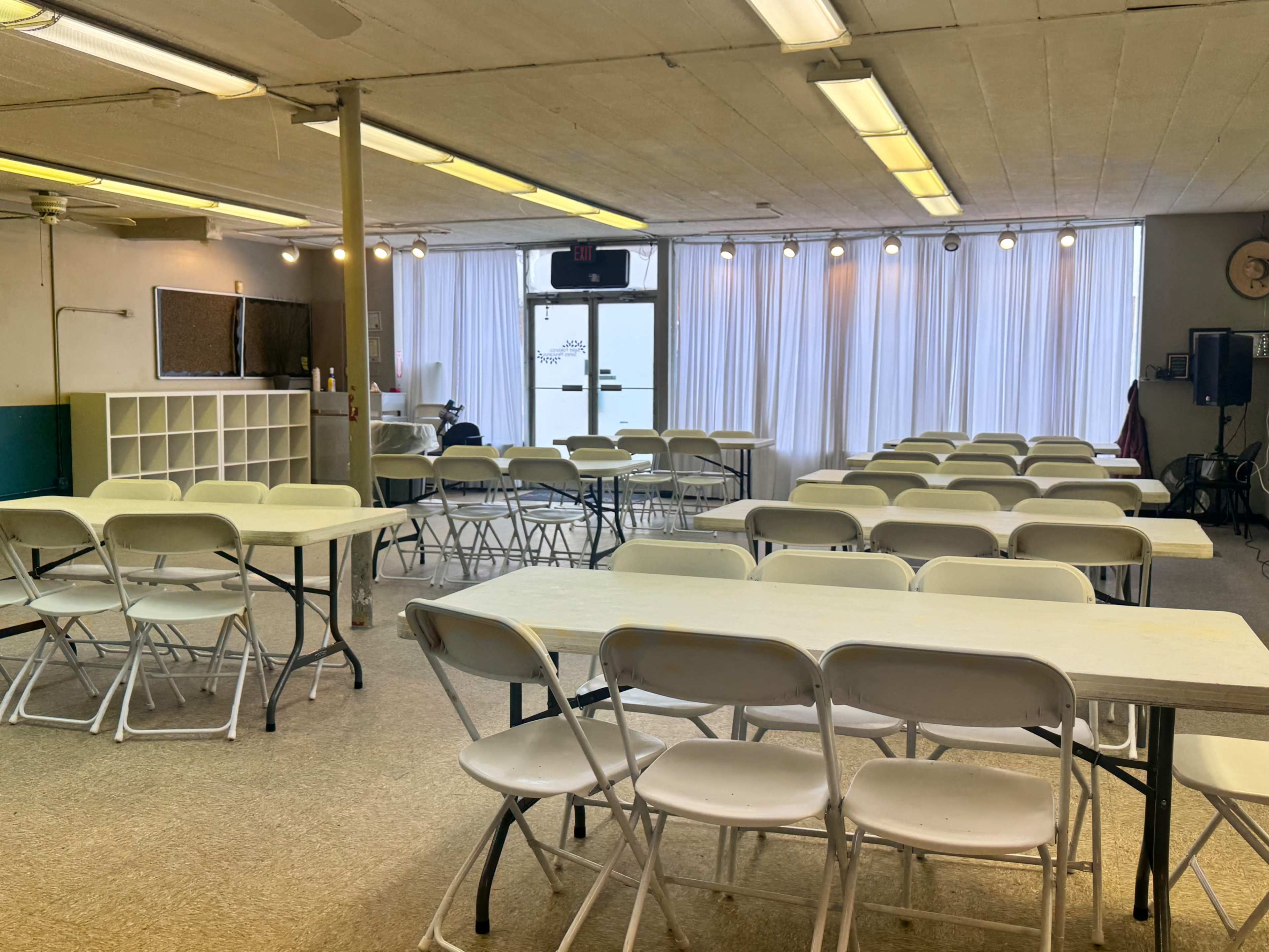 The image shows an empty room set up with several rows of folding tables and chairs, illuminated by natural light from large windows.