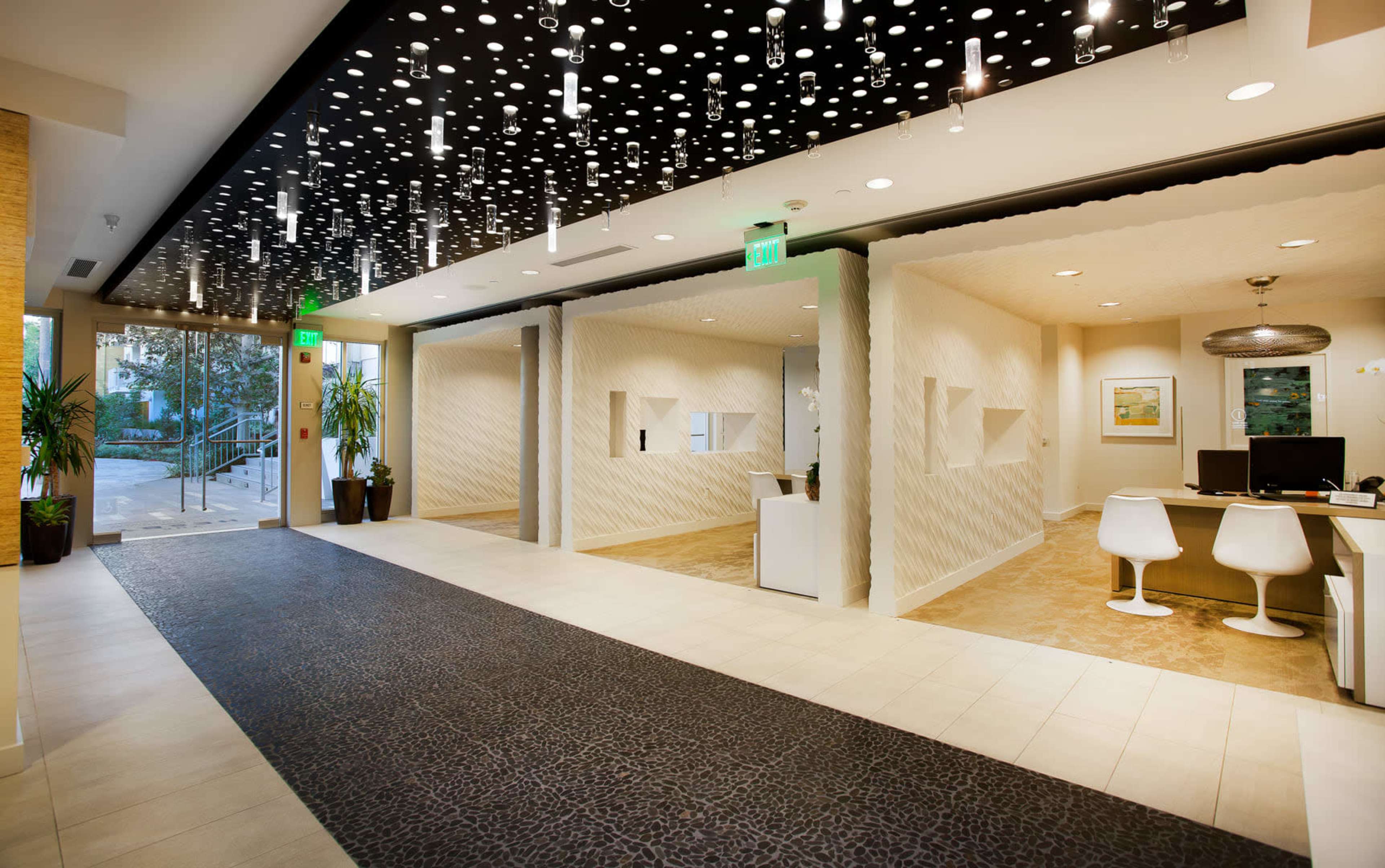 The image shows a modern reception area with a patterned ceiling, sleek white furniture, and decorative plants, leading to separate office spaces.
