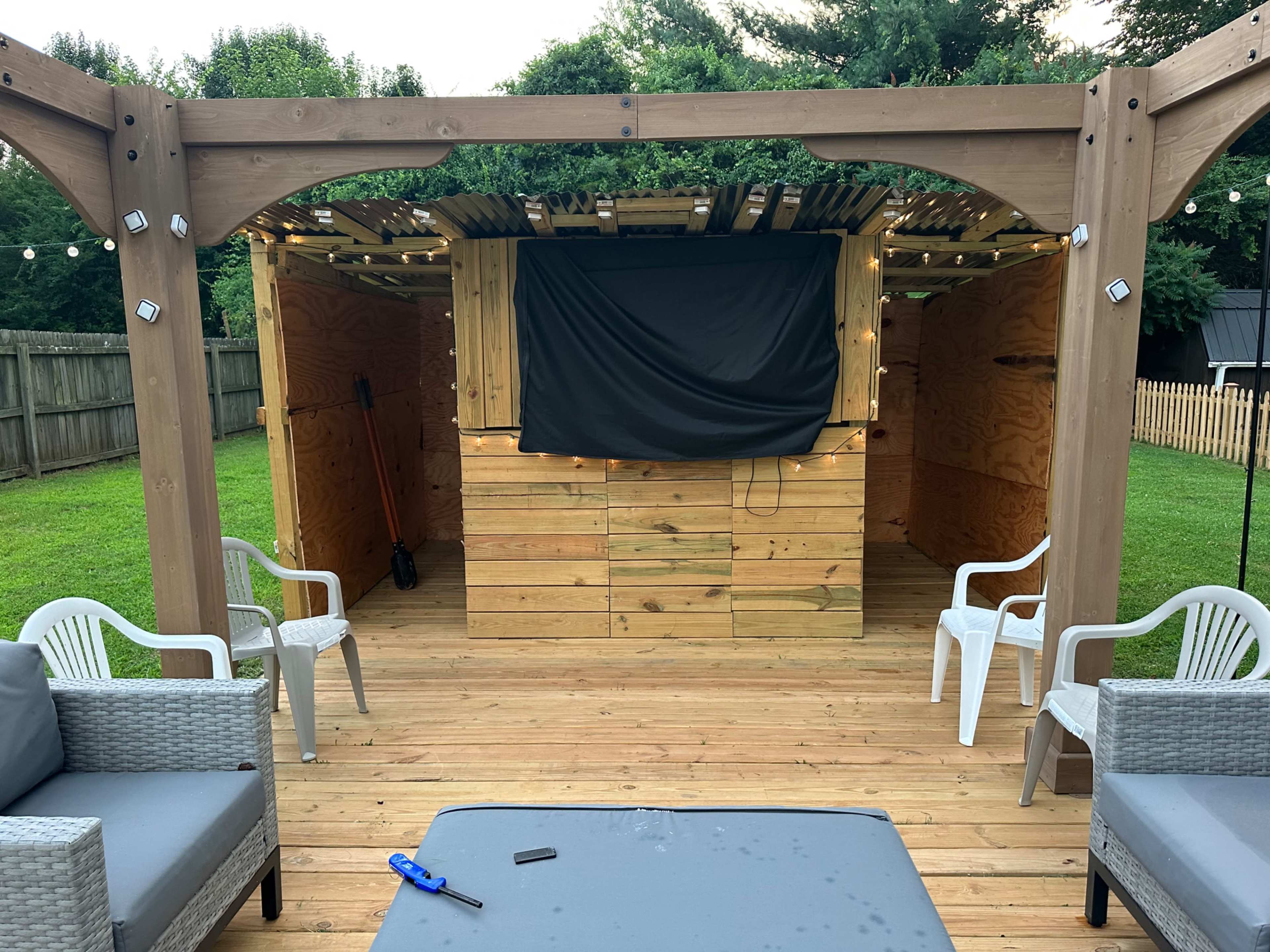 A wooden deck features a small, partially enclosed structure with a black screen and string lights, surrounded by white chairs and a table.