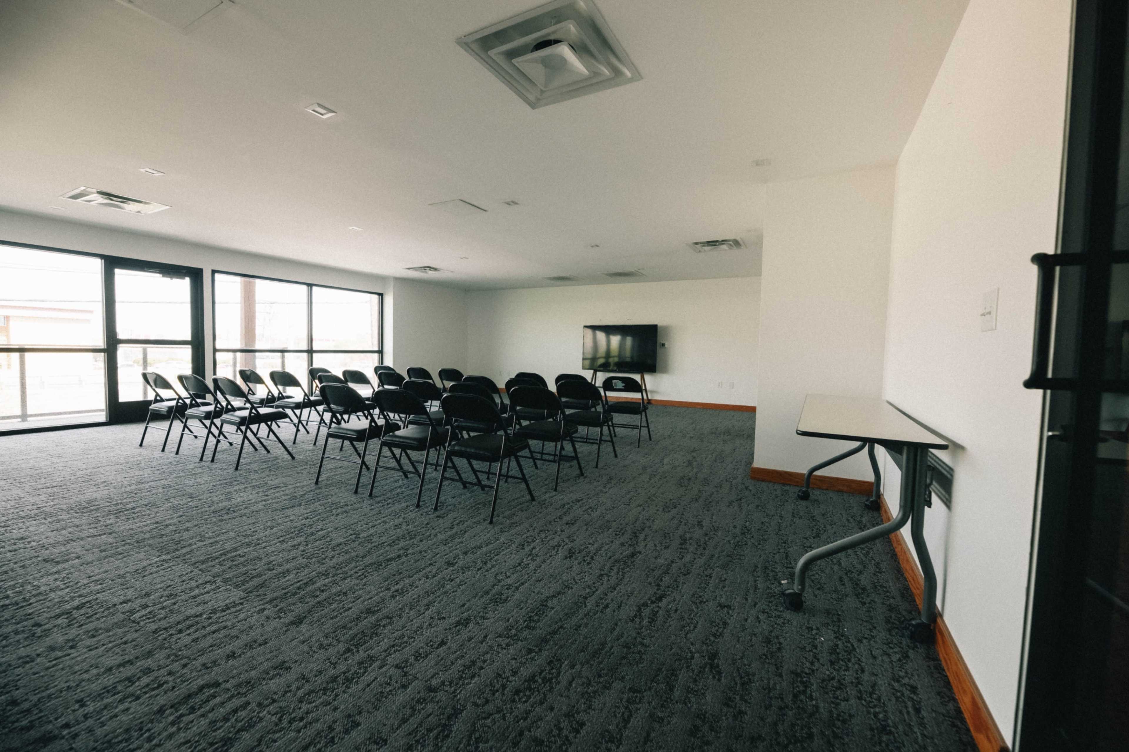 A meeting room features rows of black chairs facing a wall-mounted television, with large windows allowing natural light.