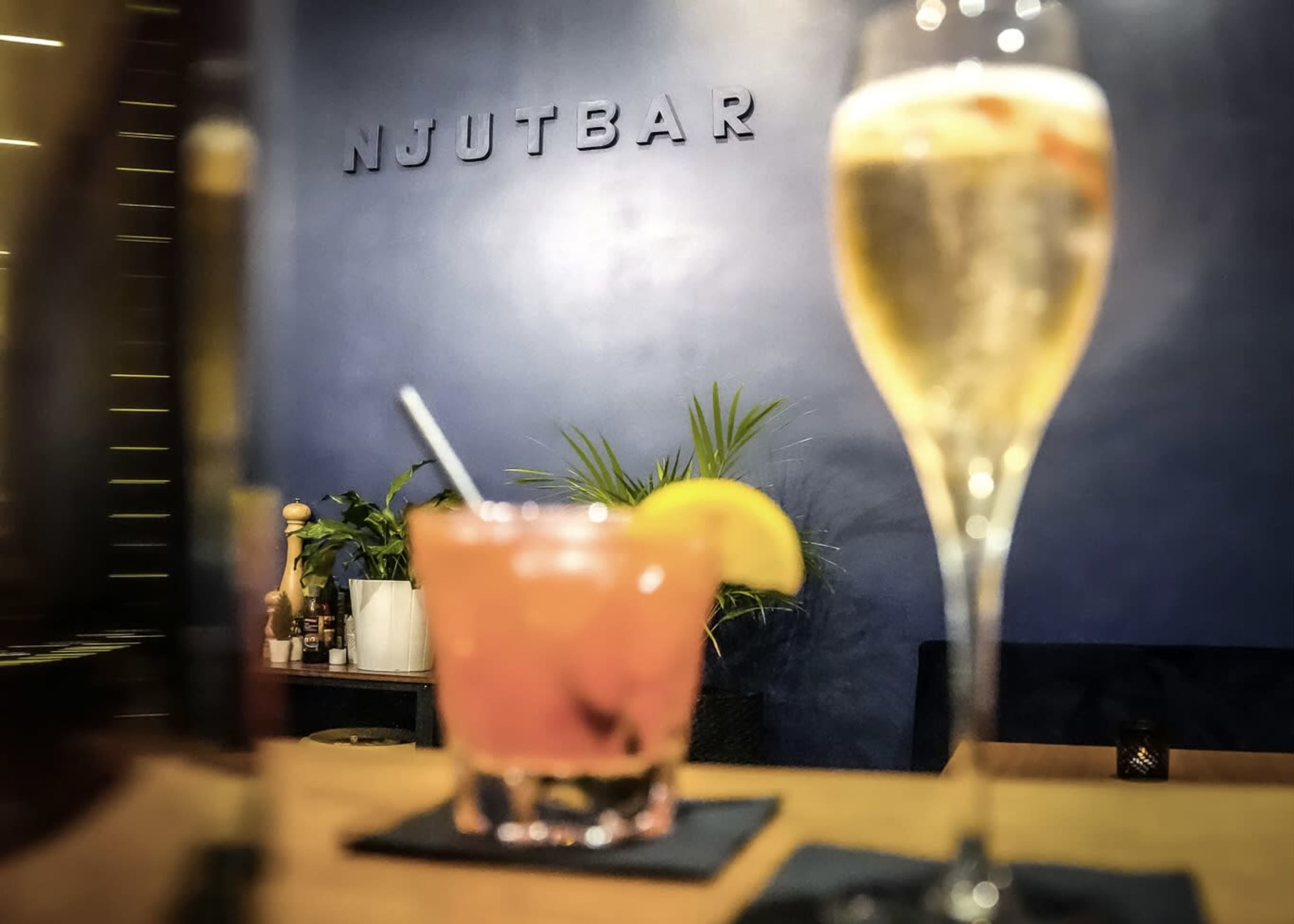 A bar counter features a cocktail and a glass of champagne with the word "NJUUTBAR" displayed on the wall in the background.