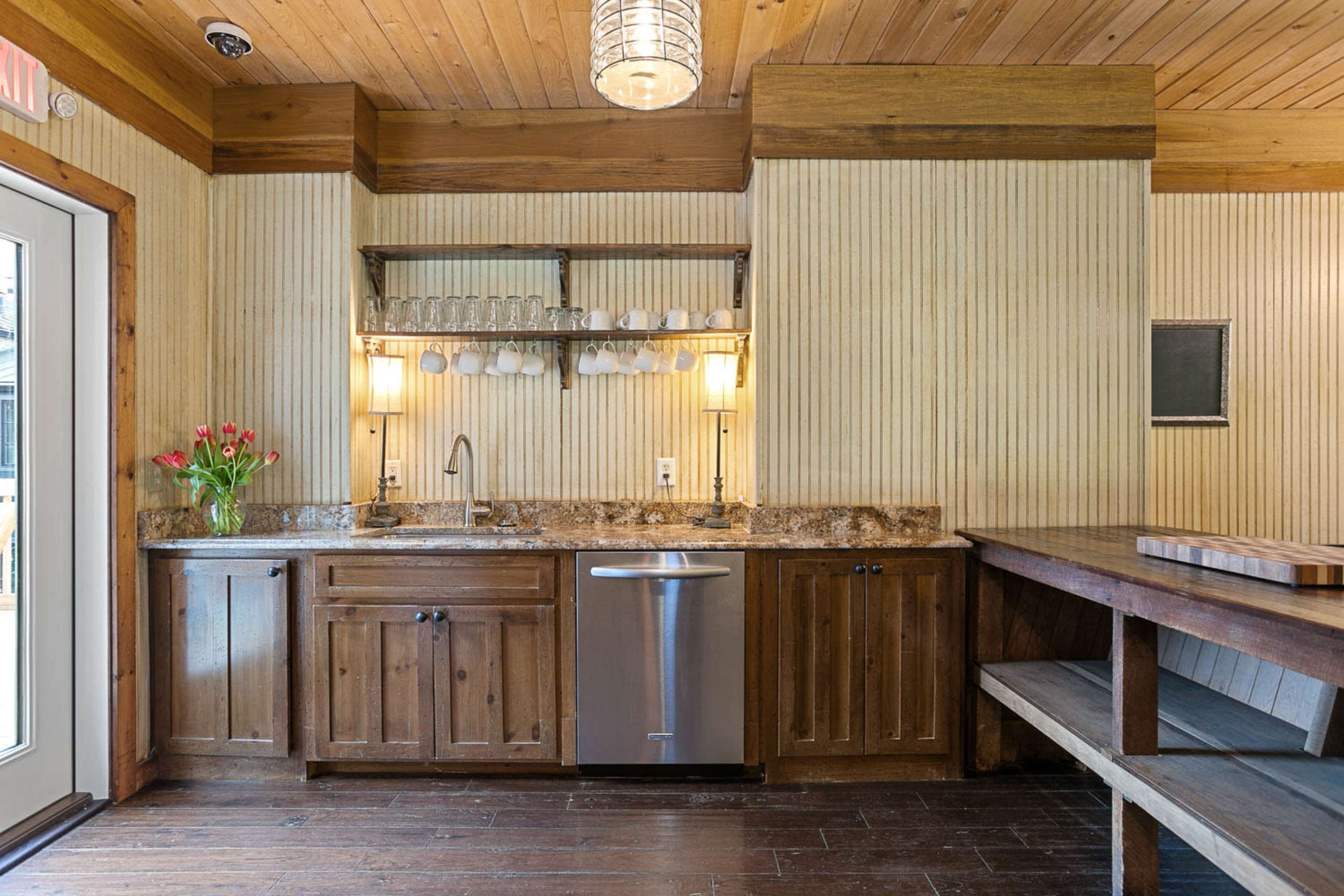 The image shows a wooden kitchen with a granite countertop, a sink, an undercounter dishwasher, and shelves displaying glasses and mugs, alongside a vase of flowers.