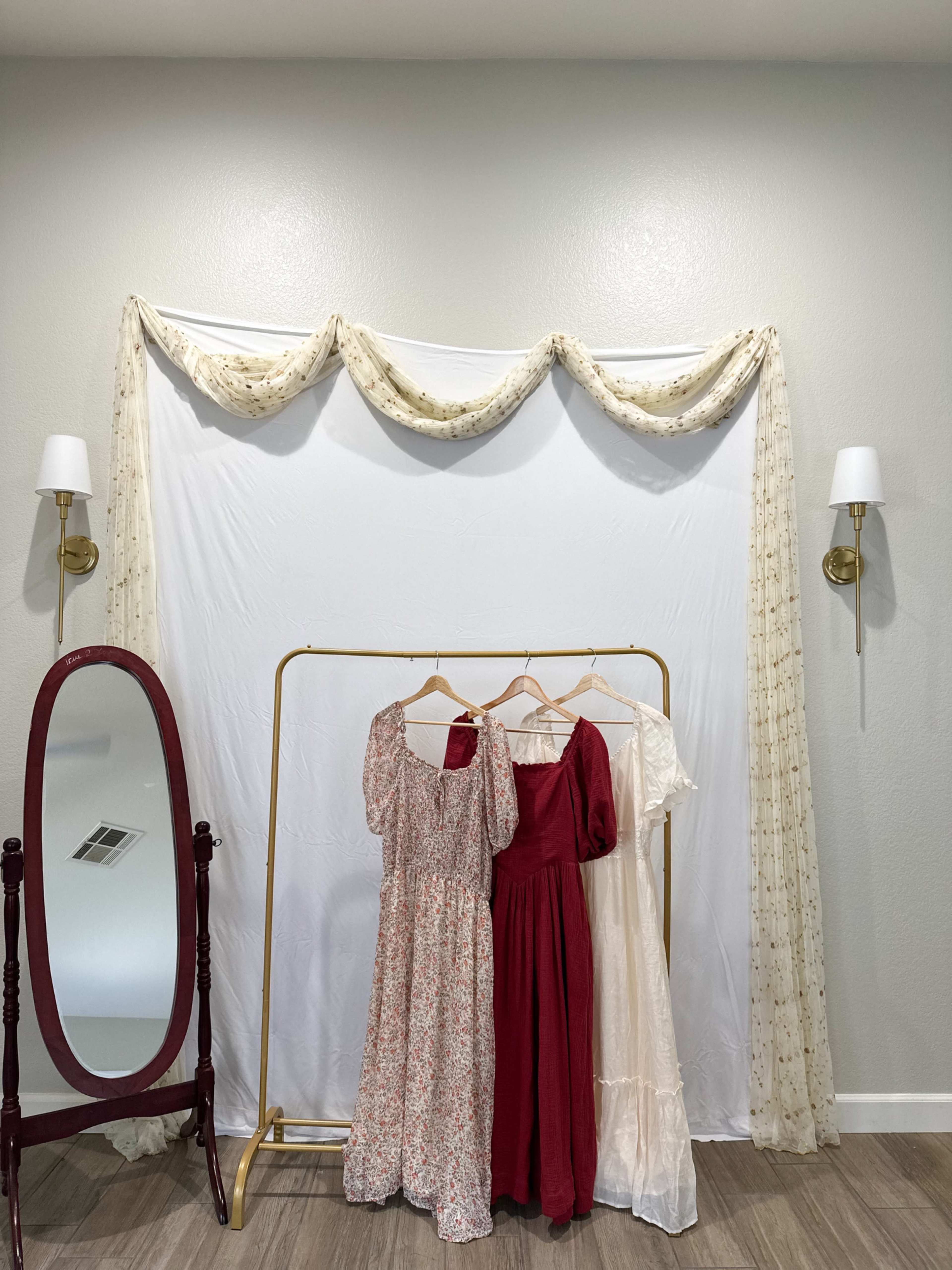 A simple dressing area features a white backdrop with draped fabric, a clothing rack displaying three dresses, and a full-length mirror nearby.