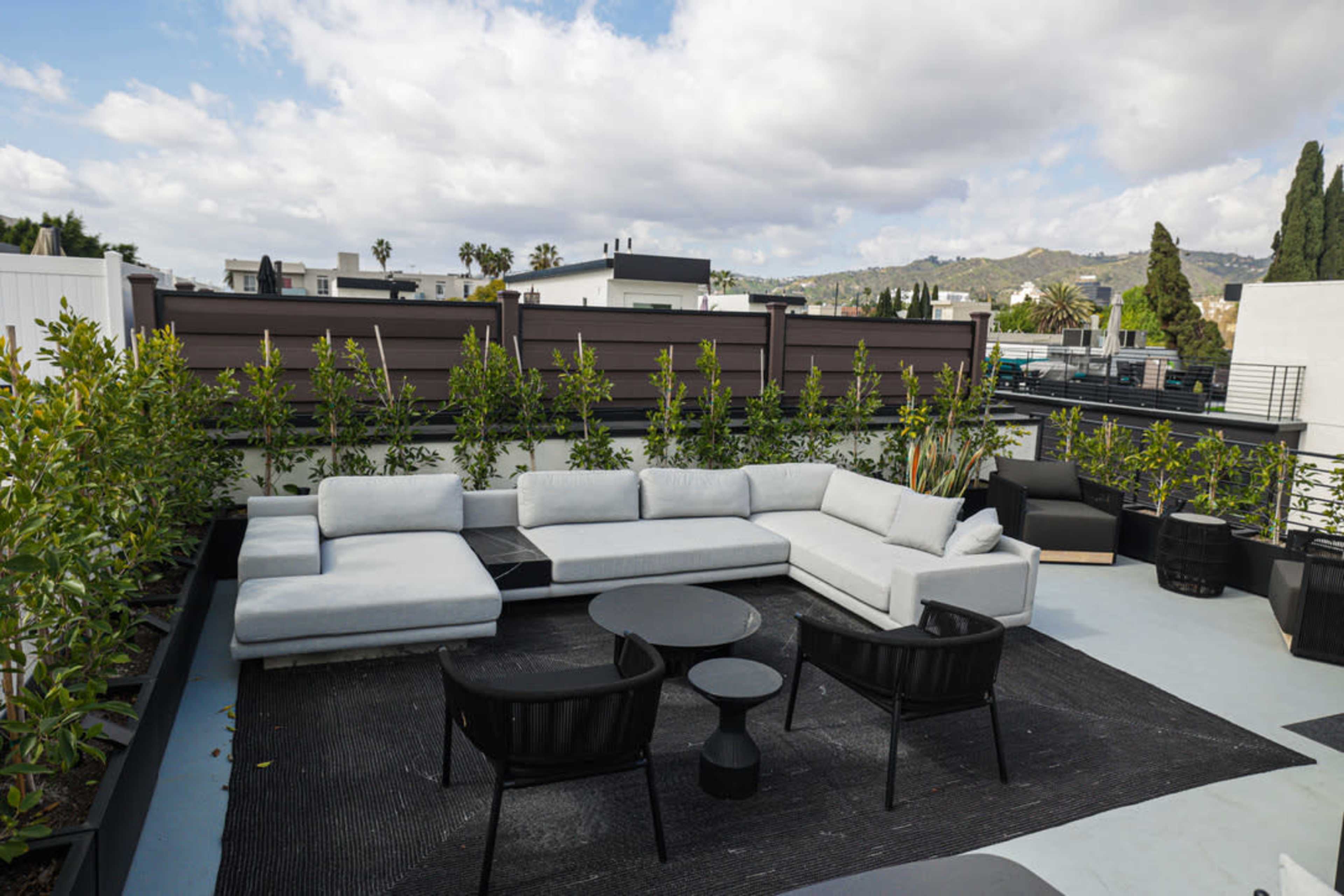 A modern rooftop lounge area featuring a sectional sofa, a circular black table, and two chairs, surrounded by greenery and a view of buildings and hills in the background.