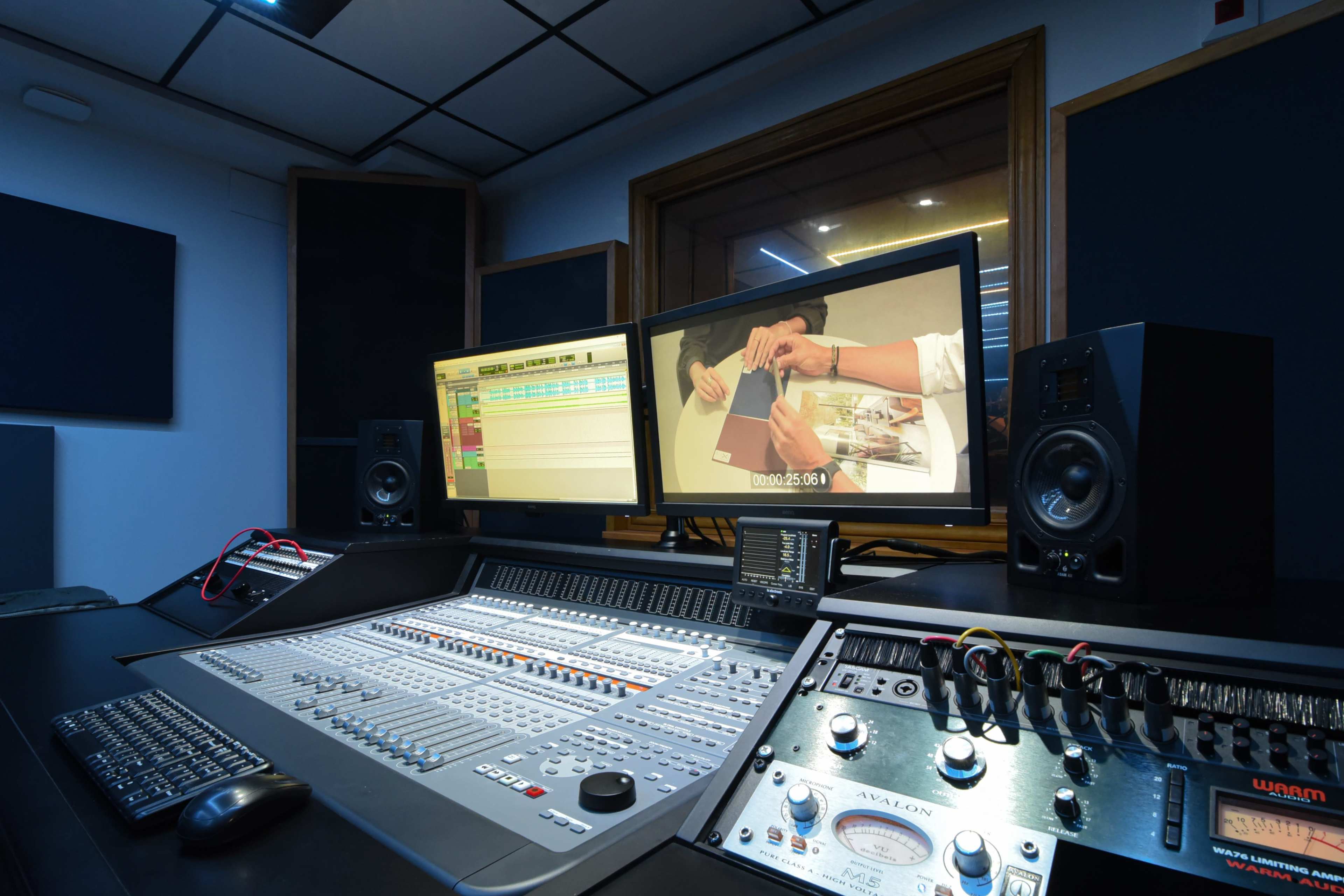 The image shows a recording studio with a mixing console, dual computer monitors displaying editing software, and speakers.