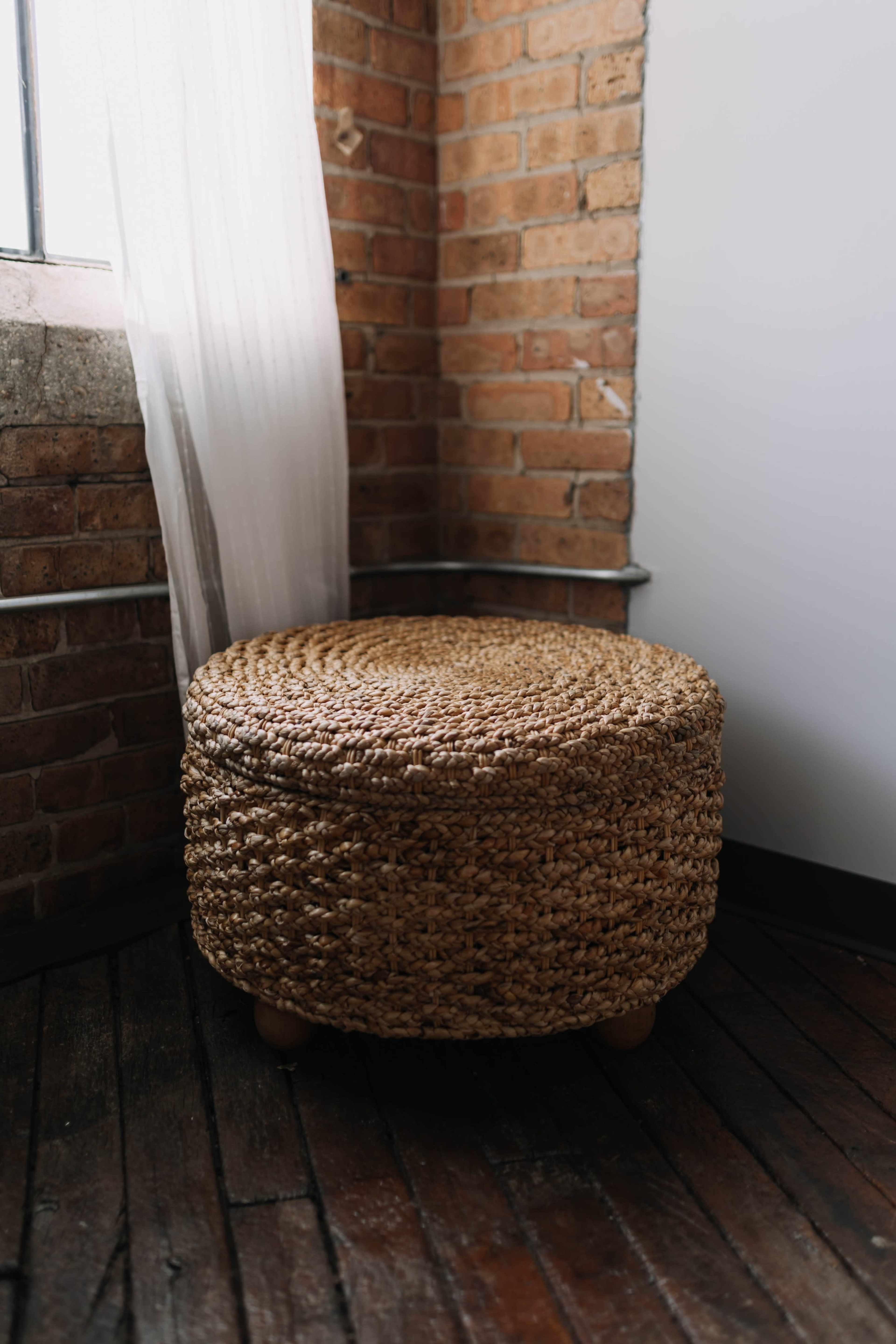 A round woven storage ottoman sits in the corner of a room with exposed brick walls and a sheer curtain.
