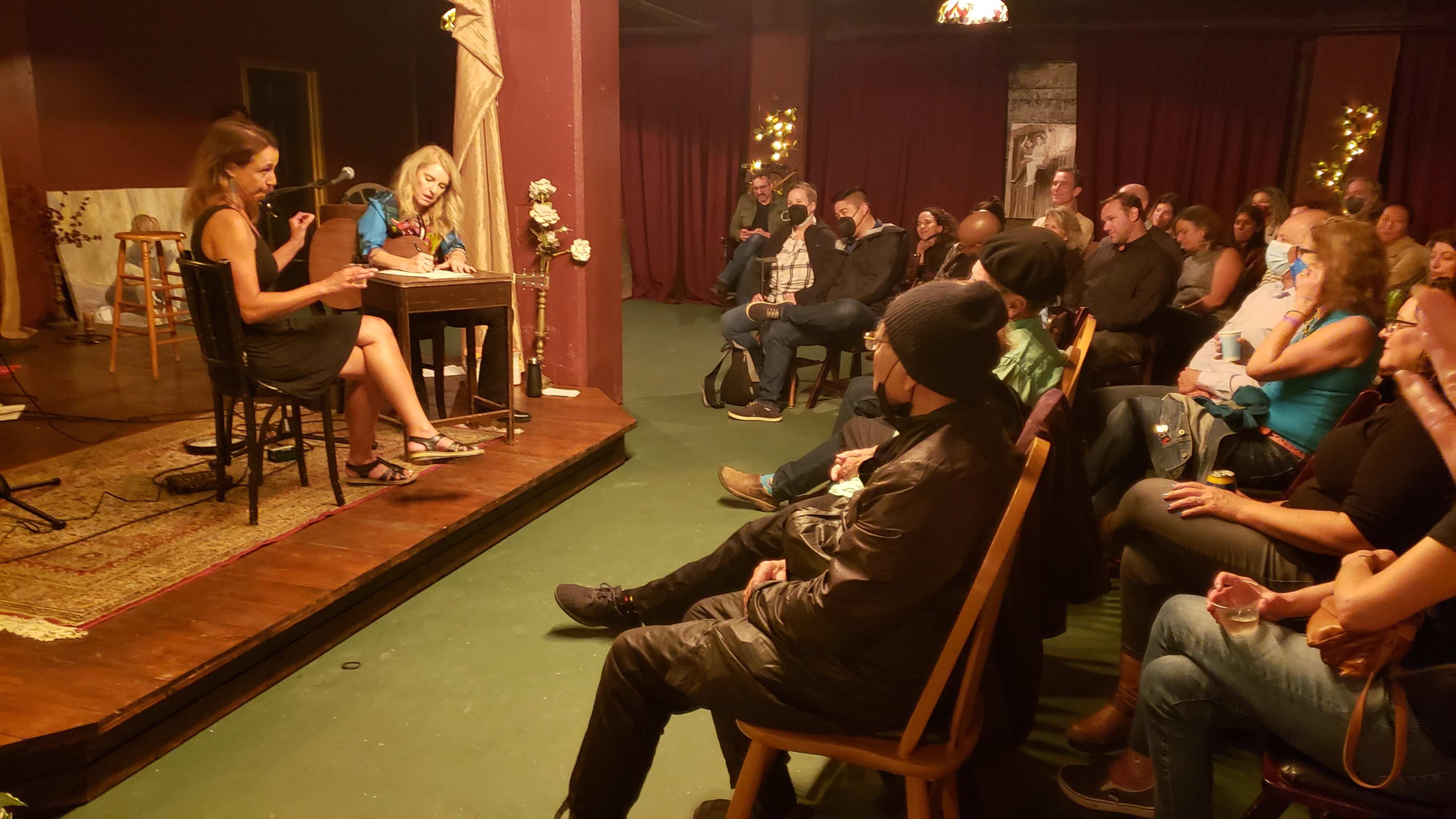 A woman reads from a script at a small venue while an audience attentively listens.