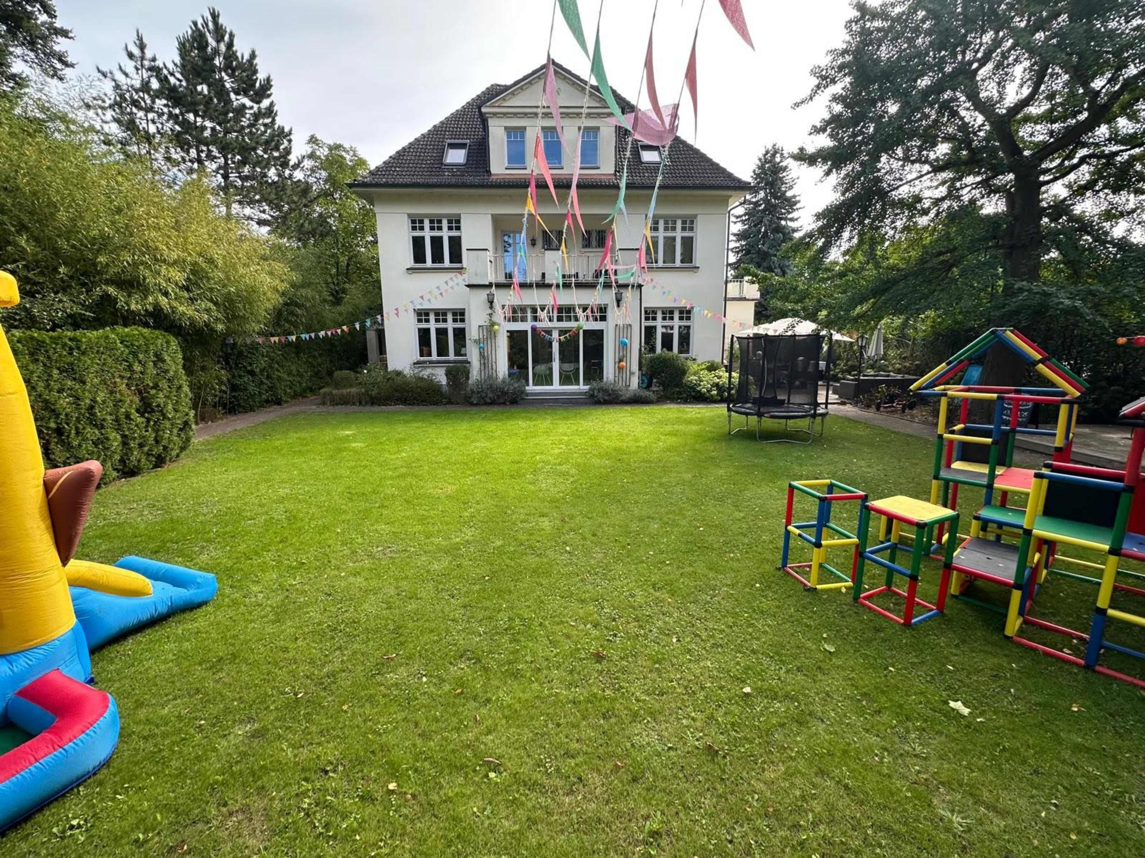 A large backyard features a house in the background, vibrant decorations, a trampoline, and colorful play structures on a well-maintained lawn.