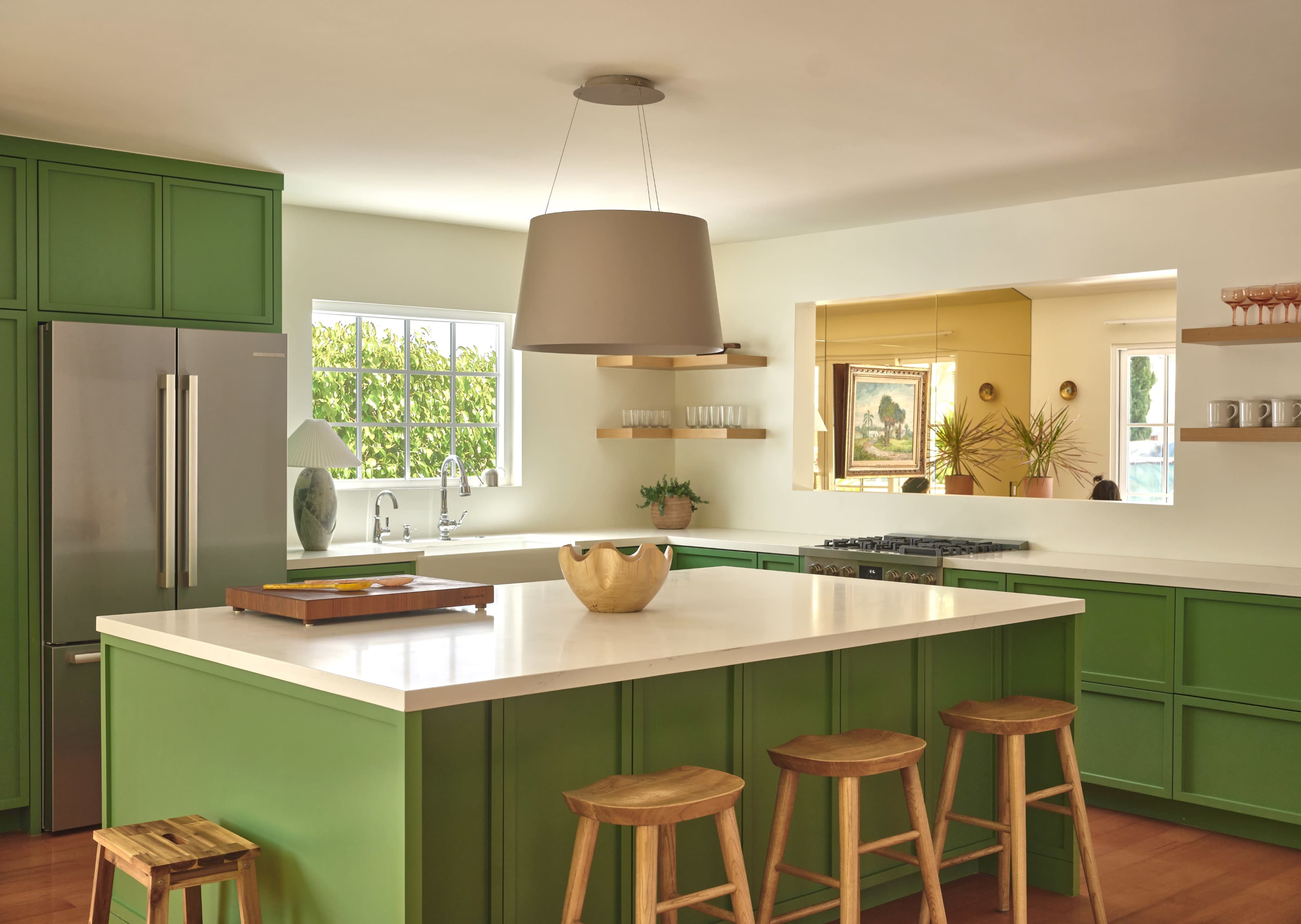 The image shows a modern kitchen with green cabinetry, a large white island, wooden stools, and a pendant light overhead.
