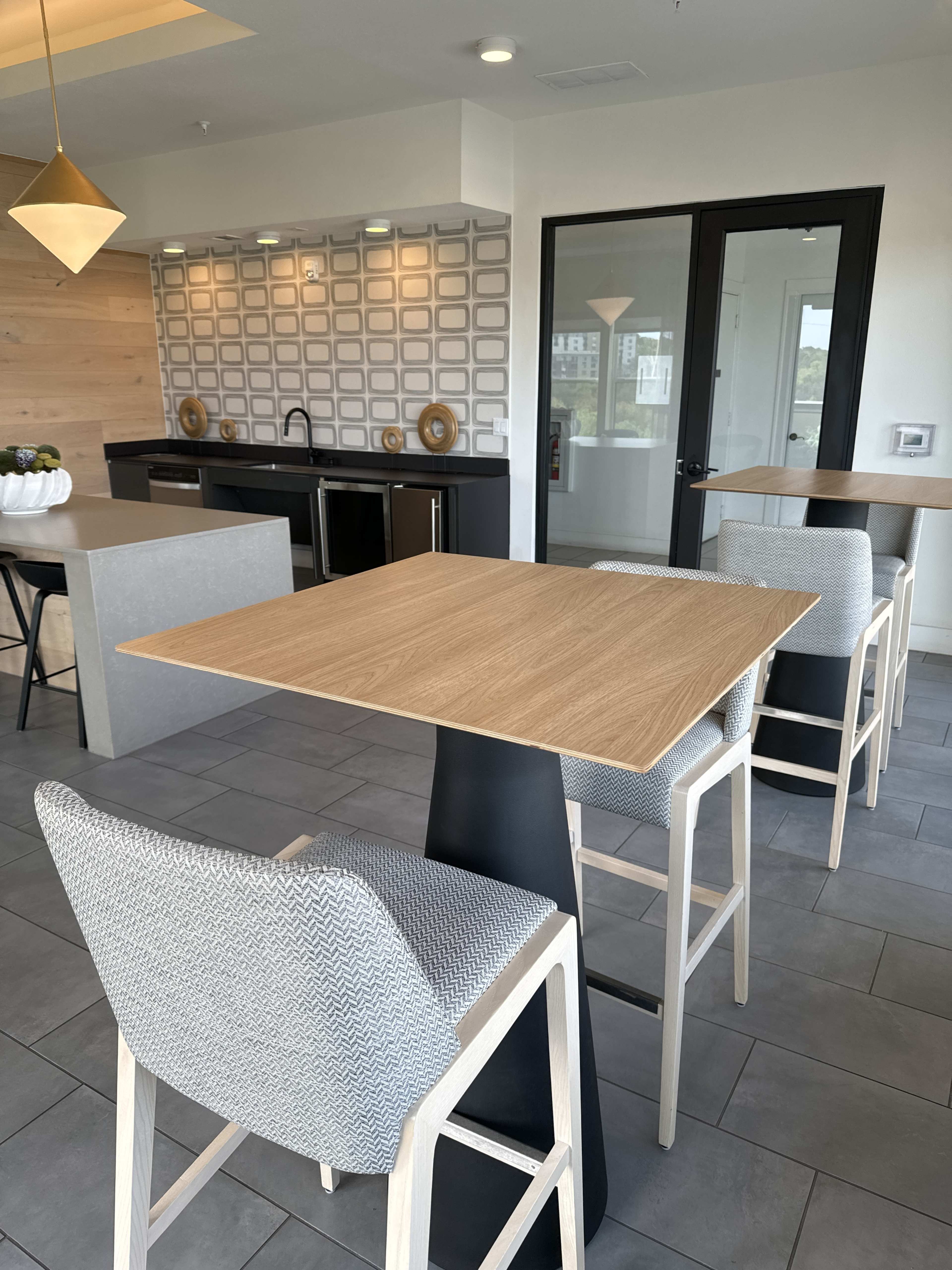 The image shows a modern kitchen area with a tall wooden table and high-backed chairs, set against a patterned backsplash and large windows.