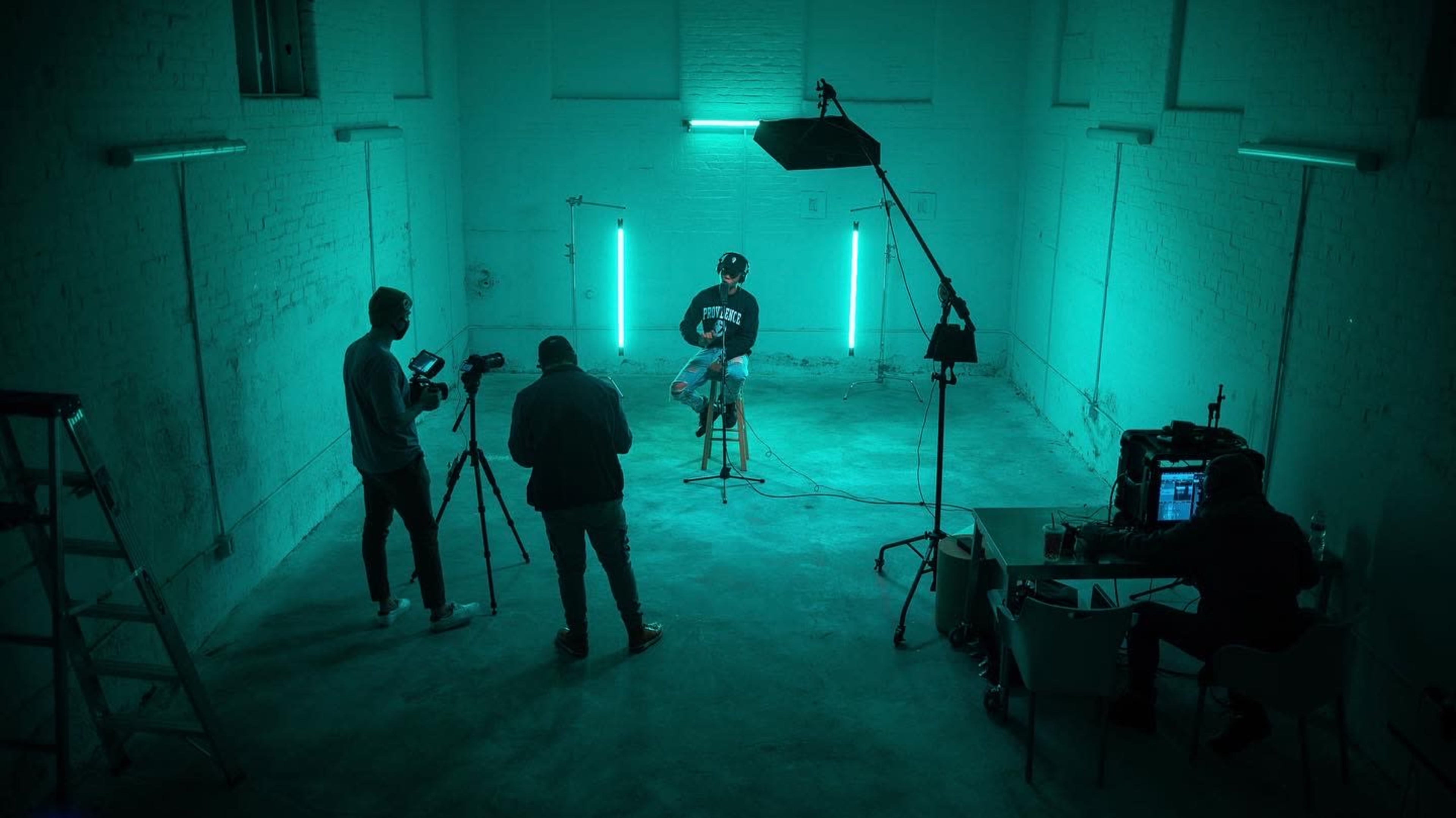 A person sits on a stool in a dimly lit room illuminated by teal lights, while two individuals operate cameras and a third monitors equipment at a desk.